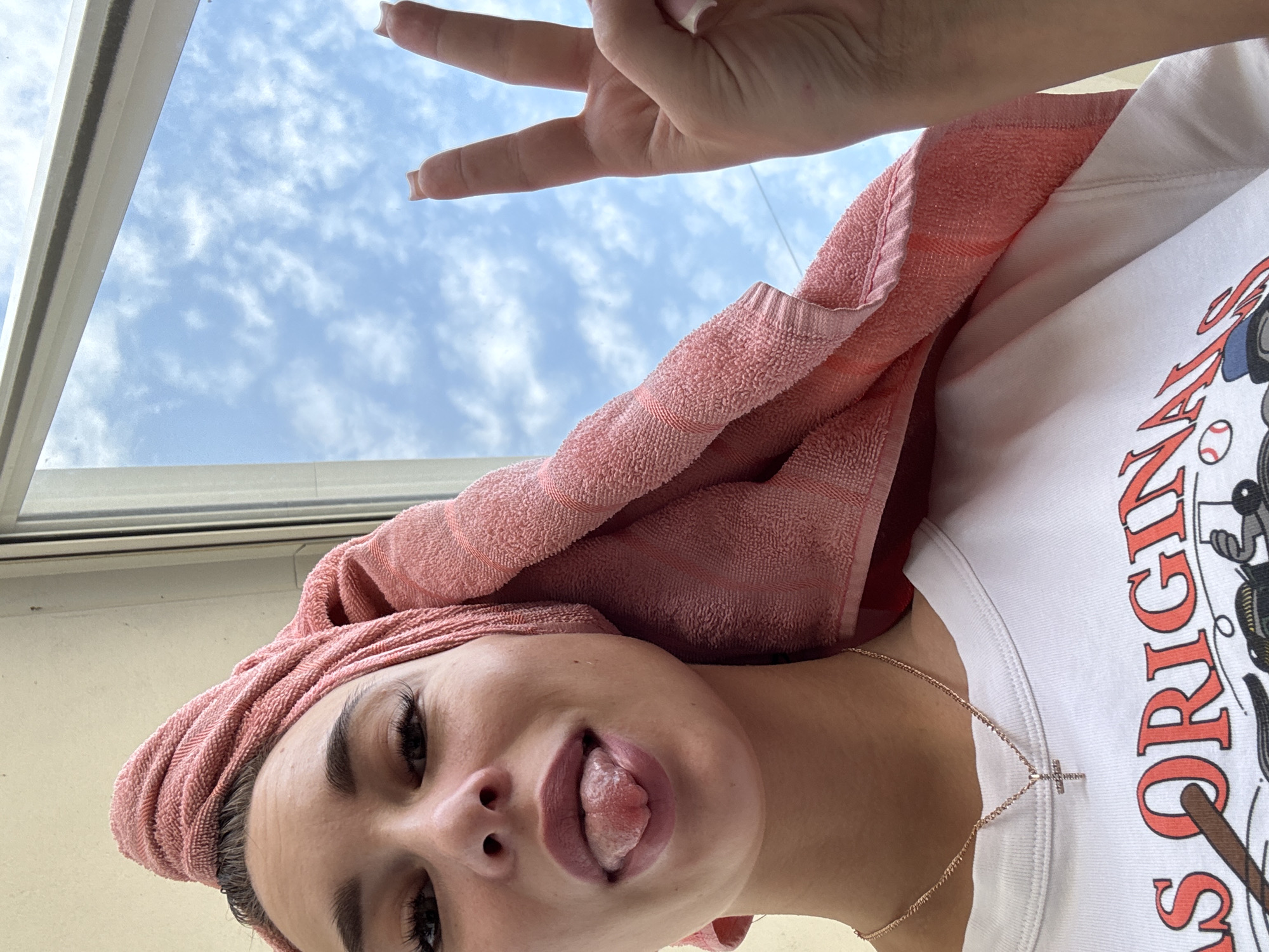 This low-angle, upside-down image captures a person, likely female, in a playful pose against a bright sky backdrop. A pink or coral towel is wrapped around her head, suggesting freshly washed hair. She is wearing a white T-shirt featuring a partial graphic and text that reads "ORIGINALS" along with what appears to be a baseball and bat motif. A thin, gold-colored chain with a small cross pendant hangs around her neck.
Her left hand is positioned in the upper portion of the frame, making a "peace sign" (V-sign) with two fingers extended, revealing light-colored fingernails. The person's face is prominent, with her tongue playfully sticking out. Notable facial features include dark, well-defined eyebrows and long, voluminous eyelashes.
The background shows a clear blue sky with scattered white, cumulus clouds, framed by a window in the upper left corner, indicating an indoor setting looking outwards. The image conveys a lighthearted and cheerful mood. The scene takes place during the daytime under fair weather conditions. The implied location is Chaikovsky, Russia.