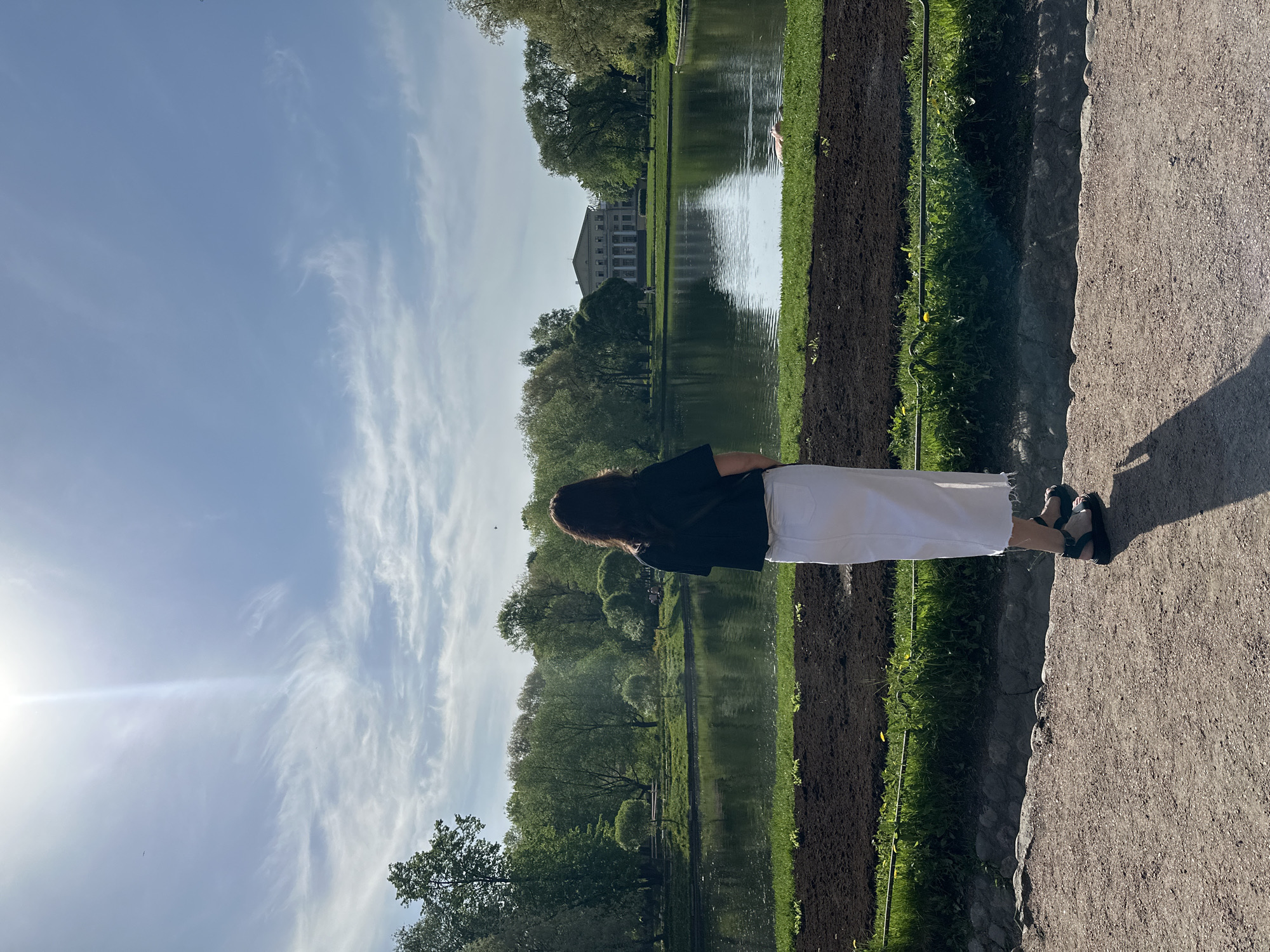 A landscape photograph taken during the day in Saint Petersburg, Russia shows a person standing on a dirt path looking out over a body of water toward a building in the distance. The person has dark hair and is wearing a black shirt and white pants. The body of water reflects the building and trees on its bank. The sky is blue with some clouds. The building in the distance is light-colored with many windows. There are trees lining the bank of the body of water. The dirt path is located on the right side of the image. There is grass and some tilled soil between the dirt path and the body of water.
