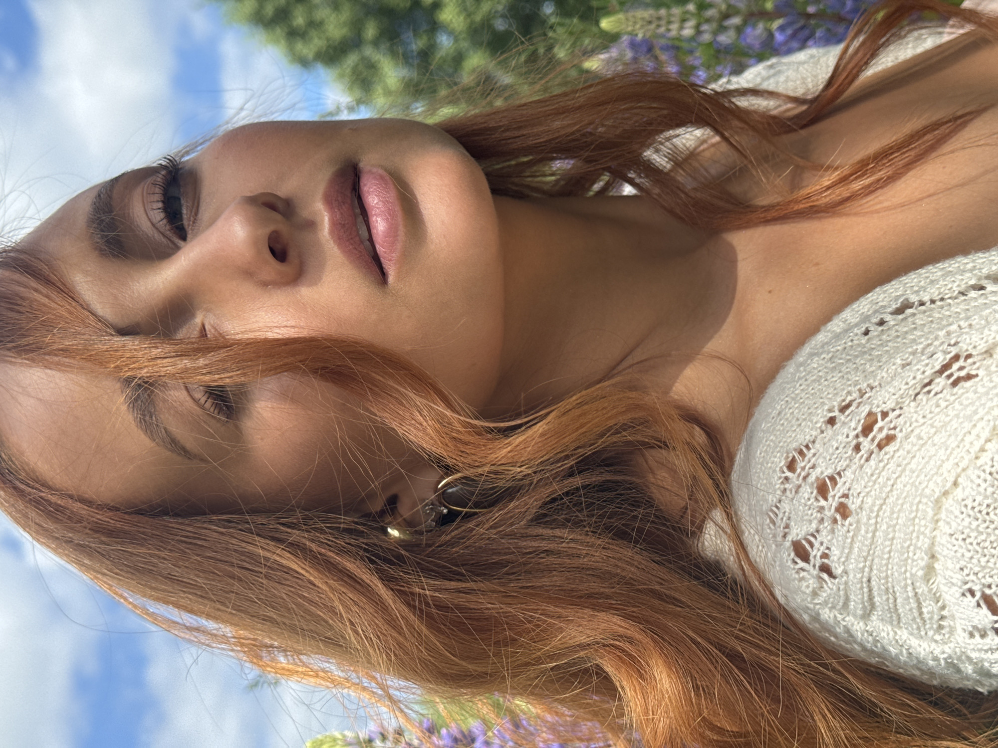 A close-up, side-profile shot captures a woman with long, auburn hair against a blurred background of lupine flowers and a blue sky with white clouds. She is wearing a cream-colored, open-knit cardigan. Her head is tilted upwards and to the right, with her eyes looking down and slightly away from the camera. Her lips are parted slightly, and she is wearing light makeup. A gold hoop earring with a dark stone is visible in her left ear. The overall lighting suggests a sunny day.