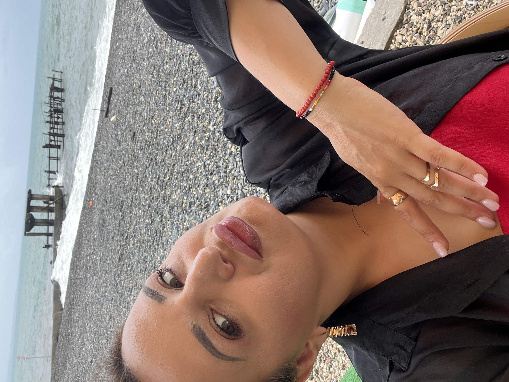 A woman is taking a selfie on a rocky beach in Sokhumi, Abkhazia. She is wearing a black shirt and a red top underneath. She has on gold earrings and two rings on her fingers, as well as a bracelet on her wrist. The ocean is in the background with waves breaking on the shore. There is also some debris in the water, possibly remnants of a pier. The sky is overcast.