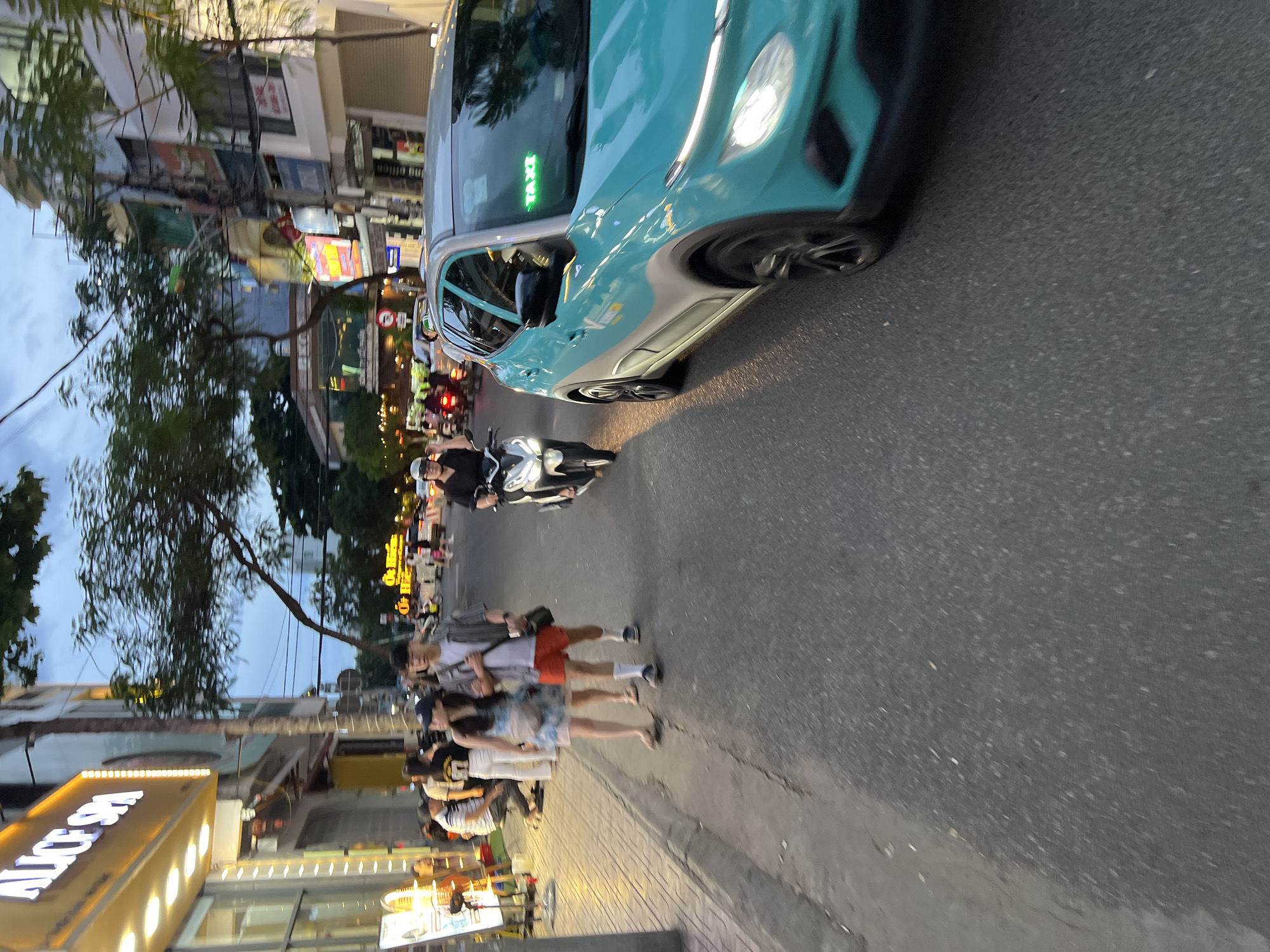The image depicts a street scene in Nha Trang, Vietnam, at dusk. A turquoise taxi with a "TAXI" sign is prominently featured on the right side of the frame, moving on the asphalt road. To the left of the taxi, a person on a white scooter is visible, also traveling on the road. Further down the street, a couple is walking on the sidewalk, the woman in a light-colored dress and the man in shorts and a striped shirt. Behind them, several other people are seen on the sidewalk and on scooters. Buildings with illuminated signs, including "ALICE SPA," line the street on the left, suggesting a commercial area. Trees with dense foliage are present along the street, and the sky is overcast with hints of twilight. The overall impression is that of a typical urban street in Vietnam during the evening hours.