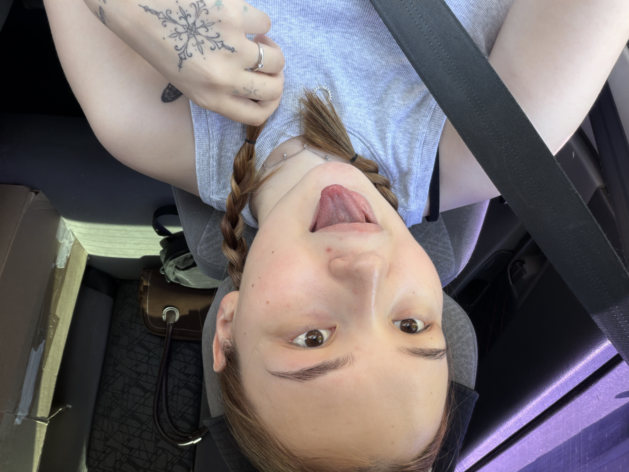 A selfie image shows a woman with light skin, brown eyes, and braided reddish-brown hair. She is sticking her tongue out. She is wearing a light gray t-shirt and is wearing a seatbelt. She has a ring on her finger and visible tattoos on her hand and arm. The setting appears to be inside a car. A box, a purse, and possibly other belongings are visible in the car. There is no verifiable information to suggest this image was taken in the city of Chaikovsky, Russia.