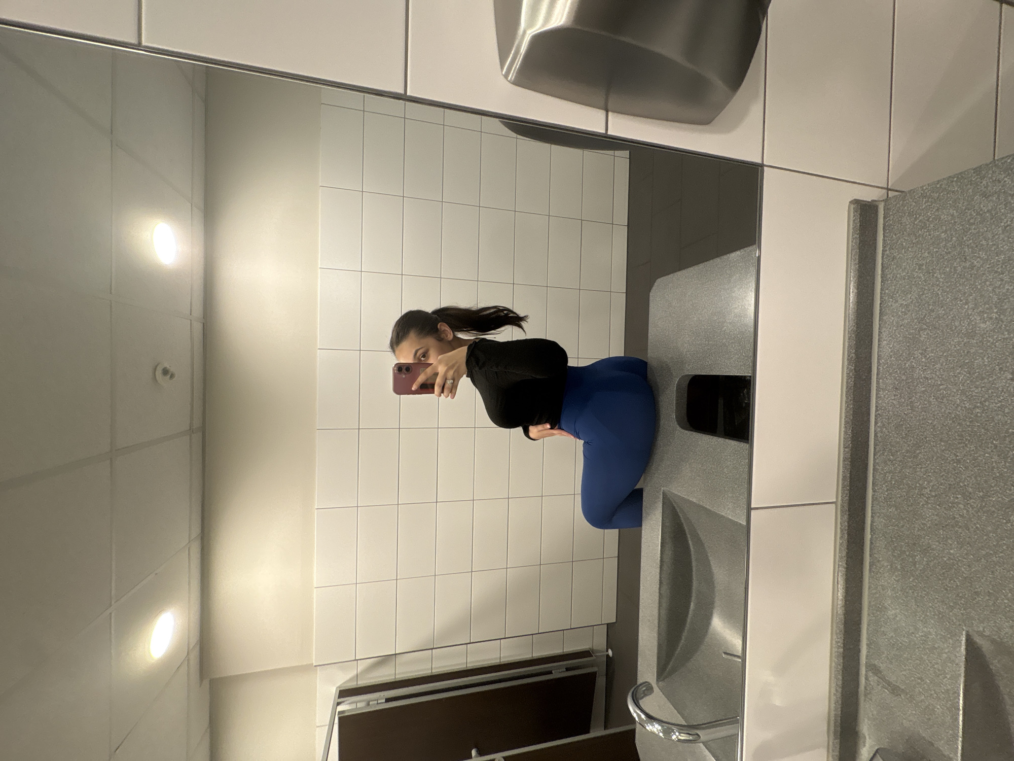 The image shows a young woman taking a mirror selfie in a public restroom. She is wearing a black long-sleeved top and blue leggings. The restroom has white tiled walls and a grey countertop with a sink. A hand dryer is visible in the reflection above the tiles. The woman is holding a pink smartphone to take the picture and is looking at her reflection. Her hair is pulled back in a ponytail. The overall setting appears to be a modern public restroom, possibly in a gym or a similar facility. There is no visible text in the image. The lighting suggests it is daytime.