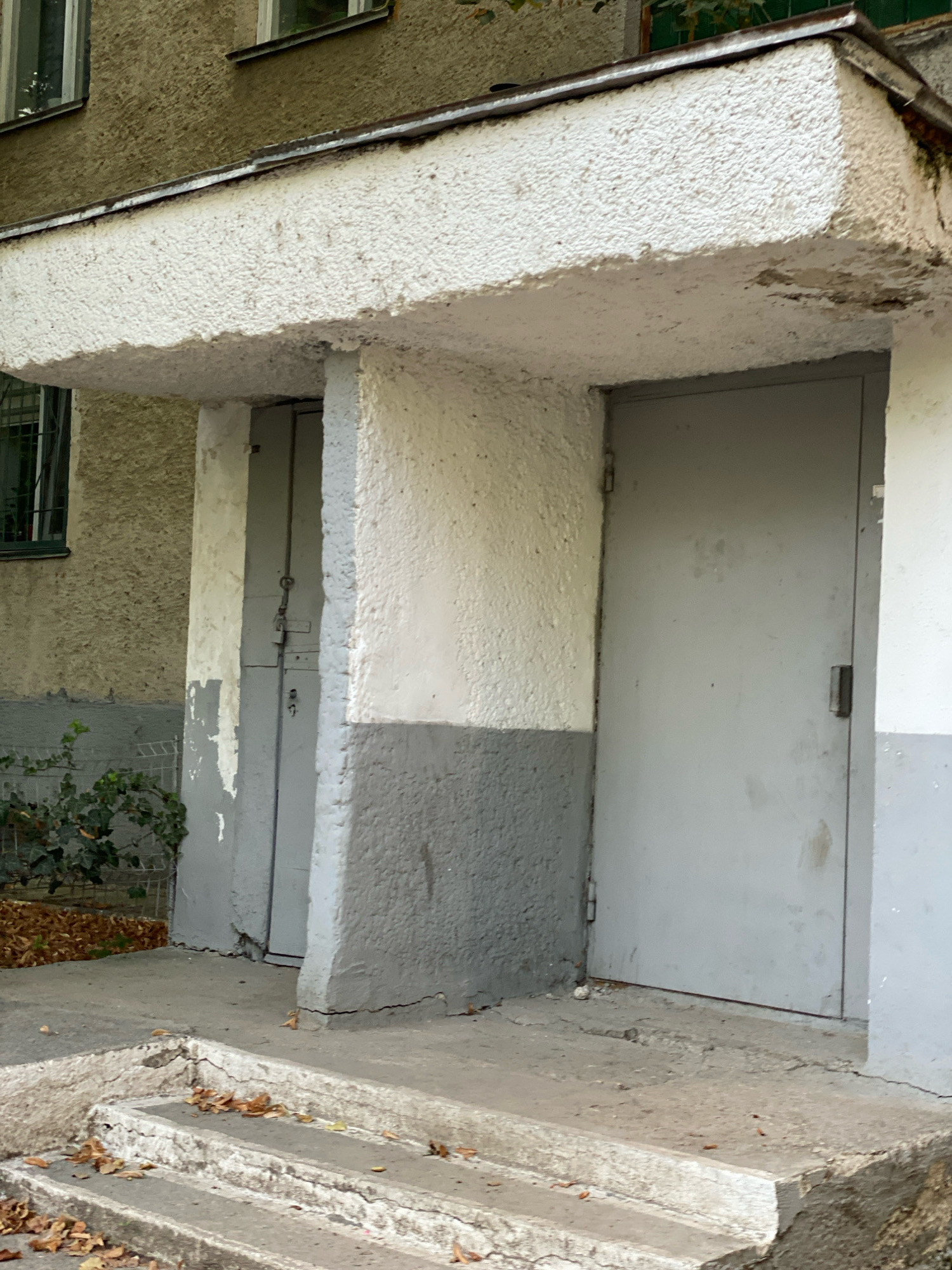 The image shows a residential building entrance in Chișinău, Moldova. The entrance is covered by a textured white overhang supported by a pillar. To the left of the entrance, there is a grey door with a padlock and what appears to be a key. To the right, a second, larger grey door is visible. The building's facade is textured and painted in shades of beige and grey. A few steps lead up to the entrance, with fallen leaves scattered on them and the surrounding pavement. Green ivy grows near the left door, and a wire fence is partially visible behind it. The overall impression is of an older, somewhat worn but functional building entrance.