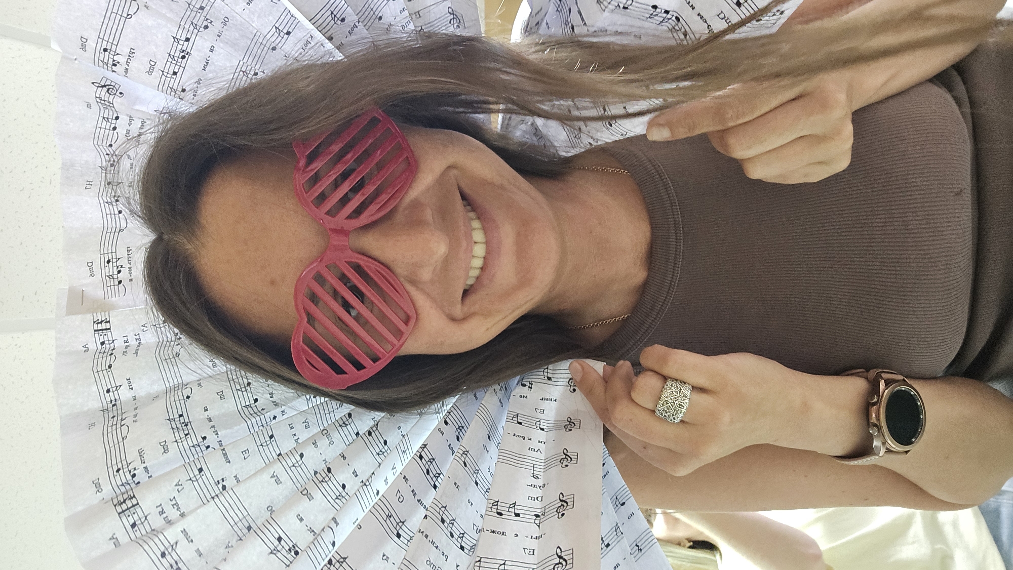 A woman is pictured in a medium shot, smiling and wearing heart-shaped pink shutter shades. She is behind a fan made of sheet music, which is fanned out behind her head and shoulders. The woman has long brown hair and is wearing a brown tank top, a silver ring, and a rose gold smartwatch. She is in an indoor environment with a plain white ceiling visible at the top of the frame.