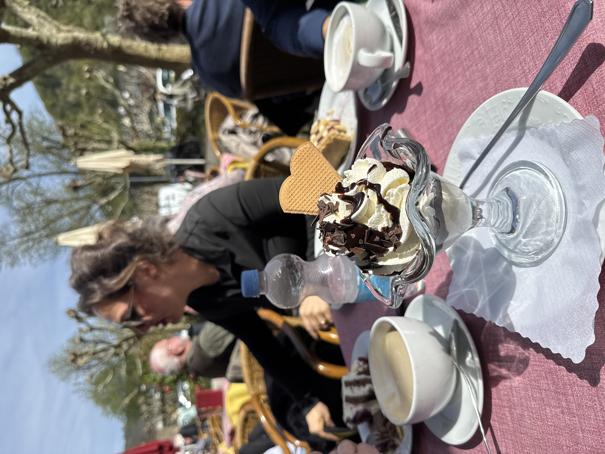 The scene depicts an outdoor café setting, likely in Cochem, Germany, given the architecture and style. The primary focus is a dessert sundae served in a stemmed glass, topped with whipped cream, chocolate syrup, and chocolate shavings, adorned with a heart-shaped waffle. The dessert sits on a white plate lined with a delicate paper doily. A long silver spoon rests on the plate. To the right of the sundae is a white cup of coffee with a saucer and another spoon. Beside it, there's a slice of cake.
A woman with sunglasses is visible in the background, seated at the table. Beside her is a water bottle. Other patrons are seated nearby, their faces partially visible. Wicker chairs are used in the café. The table is covered with a pink tablecloth. The background includes trees and possibly buildings, indicating the café's location in an urban or semi-urban environment. The time of day is likely daytime, suggested by the daylight and shadows.