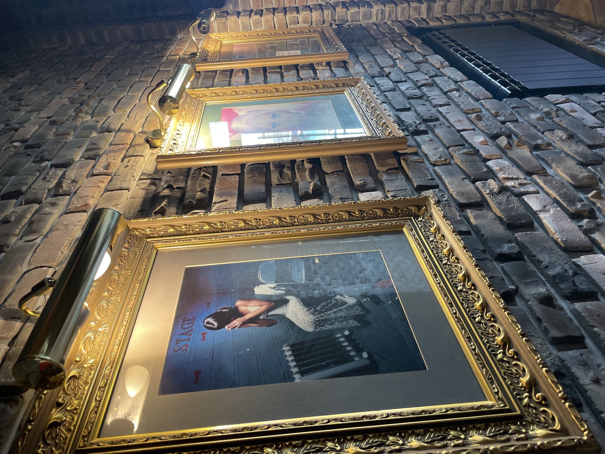 Here's a detailed description of the image:
**General Scene:**
The image shows a brick wall with three framed pictures hanging on it. A metallic lamp illuminates each picture. The perspective is from below, looking upwards at an angle.
**Objects:**
* **Frames:** Three framed pictures with ornate golden frames.
* **Pictures:** The bottom picture contains the word "Stage" in bold letters. The middle picture appears to be a photo of a woman. The top picture is difficult to discern from the low angle.
* **Lamps:** Metallic lamps are mounted next to each frame, directing light onto the pictures.
* **Window:** A window is at the top right of the frame.
**Environment:**
* **Wall:** The pictures and lamps are mounted on a brick wall. The bricks are various shades of brown and gray.
**Location:**
The exact location in Krasnoyarsk, Russia, cannot be determined based on the information in the picture alone.