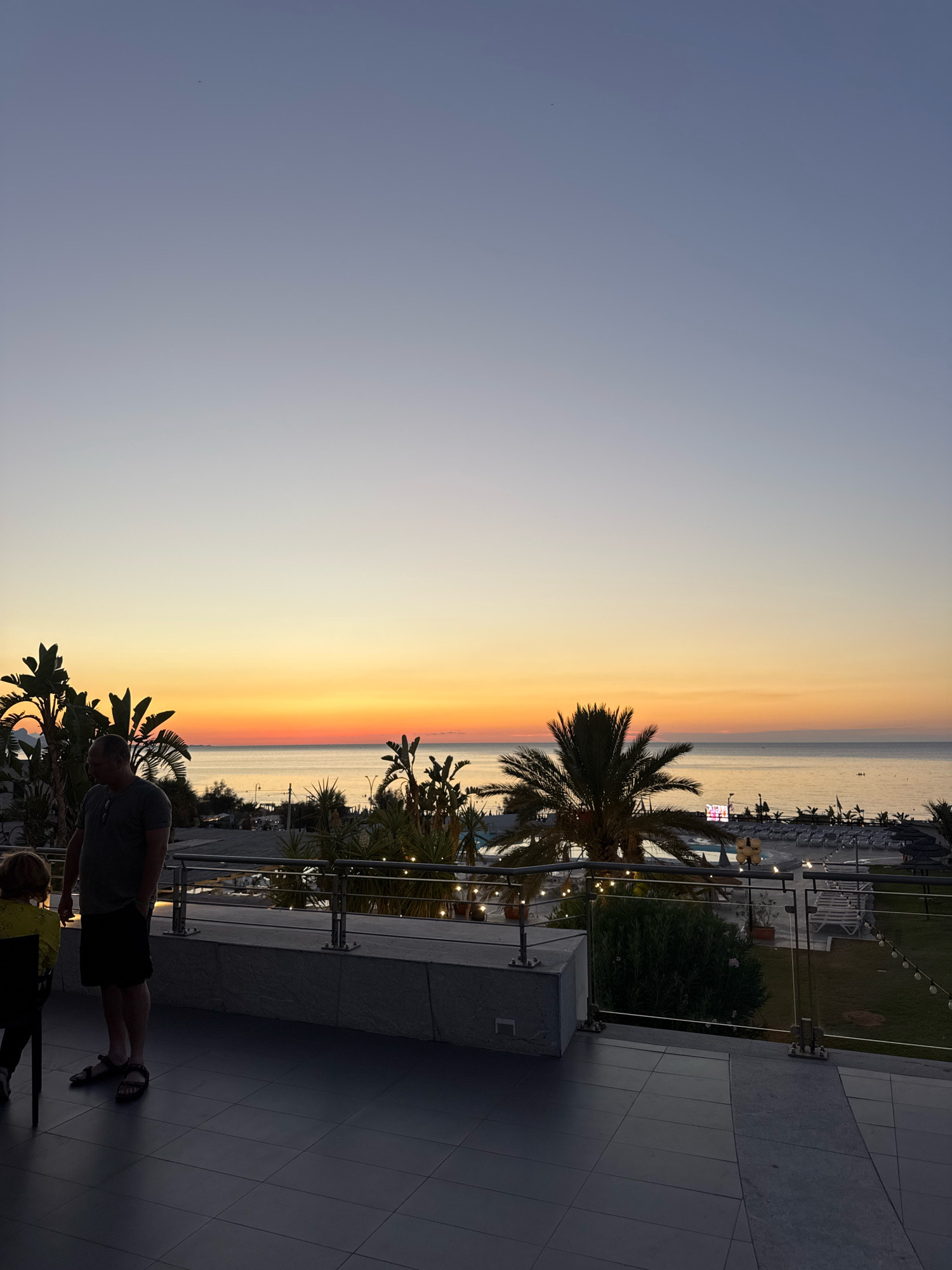 The image captures a serene sunset scene at a coastal location, likely Isola delle Femmine, Italy. A man and a child stand on a tiled balcony, overlooking a beach area with lounge chairs and umbrellas. Lush green foliage and palm trees dot the landscape, with string lights adding a warm ambiance. The sky transitions from a pale blue at the top to vibrant shades of orange and yellow near the horizon, where the sea meets the sky. The overall impression is one of peaceful relaxation and natural beauty.