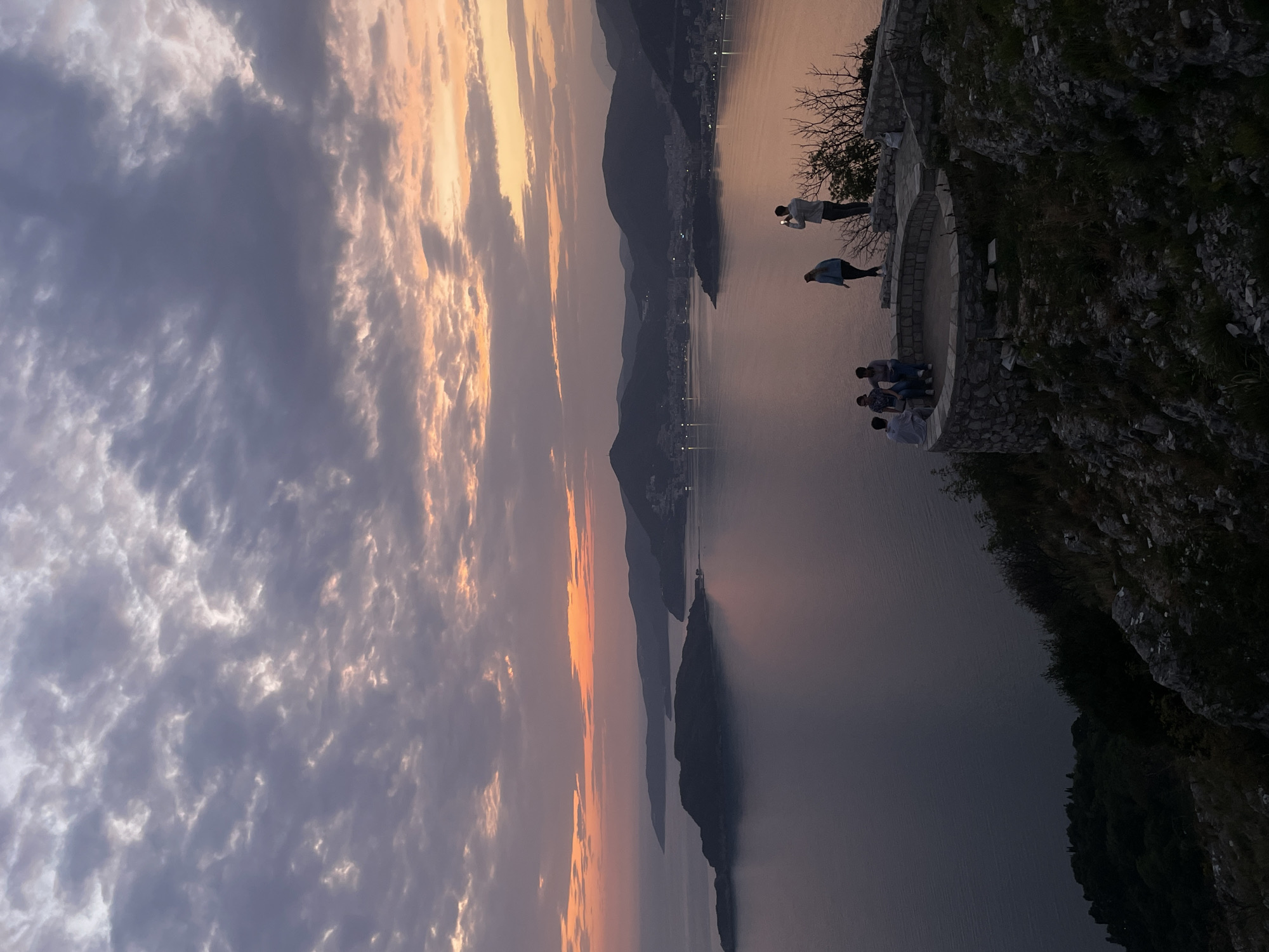The image depicts a breathtaking panoramic view of a coastal landscape at either sunrise or sunset, with the provided context identifying the location as Sveti Stefan, Montenegro. The sky is a dramatic canvas of clouds, transitioning from warm orange, pink, and purple hues near the horizon, where the sun is either rising or setting, to cooler grey and blue tones overhead. Scattered clouds add texture and depth to the vast expanse.
In the foreground, a rugged, dark cliff face, partially covered in sparse vegetation, hosts a stone viewing platform. Several individuals are present on this overlook, appearing to admire the expansive vista. To the left, two people stand near the edge of the platform; one, dressed in a light shirt, is holding a smartphone, likely photographing the scenic view. Closer to the cliff face, a small group of three to four people are gathered, also gazing out over the water.
The middle ground features a large, calm body of water, possibly the Adriatic Sea, reflecting the muted colors of the sky. Dark, mountainous landmasses or islands rise majestically from the water, silhouetted against the colorful horizon. Along the distant shores, subtle lights indicate the presence of towns or settlements. The overall atmosphere is serene and appreciative of the natural beauty of the Montenegrin coastline. No visible text is present in the image.