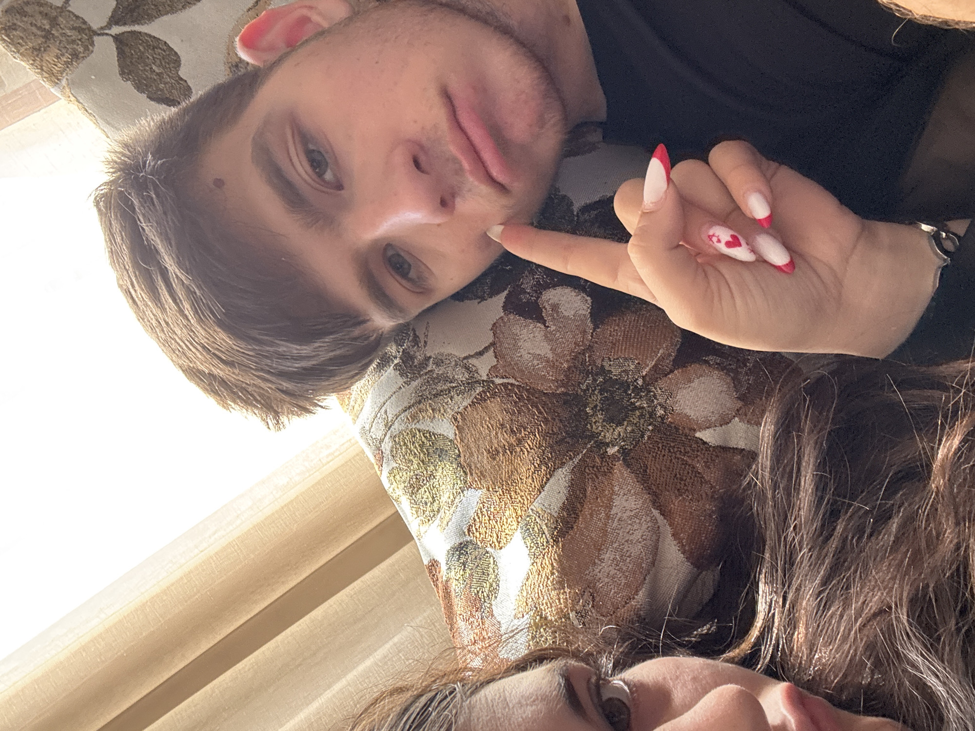 A man and a woman are lying down. The man has short brown hair, light eyes, and a slight stubble. A woman's hand with red and white manicured nails is touching his cheek. The woman has long, dark hair. They are lying on a patterned pillow. The scene is near a window with light-colored curtains. There is no evidence of a specific location in Plovdiv, Bulgaria.