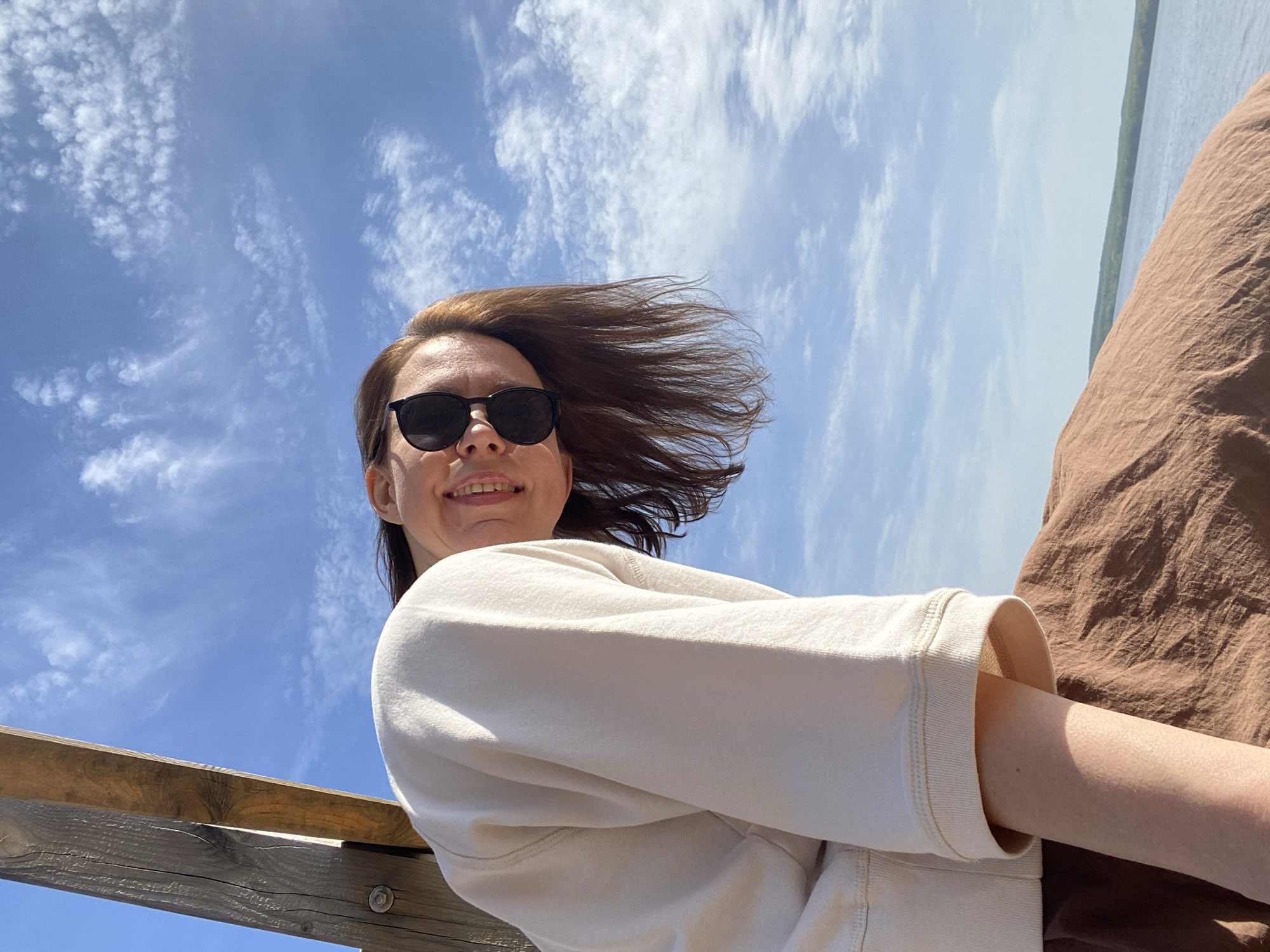A young woman with brown hair is posing for a photograph outdoors, likely on a sunny day in Chaikovsky, Russia. She's wearing sunglasses and a light-colored sweatshirt. Her hair is blowing in the wind. In the background, there is a blue sky with scattered clouds and a glimpse of a body of water lined with trees. A wooden railing is visible in the lower left corner of the frame. Part of a brown fabric, perhaps clothing, is present on the right side of the frame.