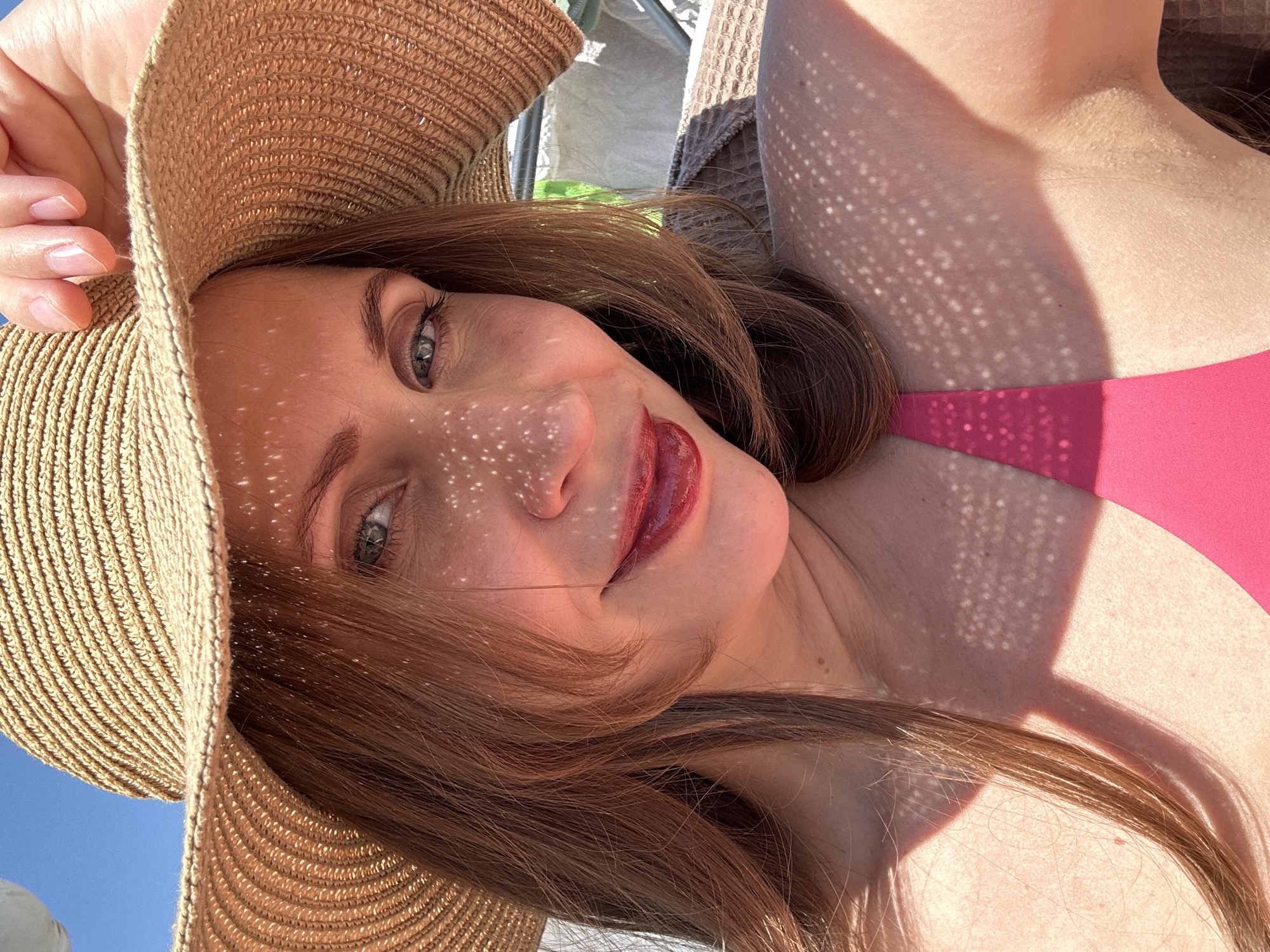 A woman with auburn hair and a straw sunhat is seen in a selfie-style shot. The woman wears a pink bikini top. The light is bright, creating speckled shadows on her face and body, possibly from the hat. The background is somewhat blurry, showing hints of a bright blue sky, white fabrics, and some green vegetation. The location can be inferred to be outdoors in Fethiye, Turkey, given the attire, sunny weather, and potential beach-like or resort-like setting.