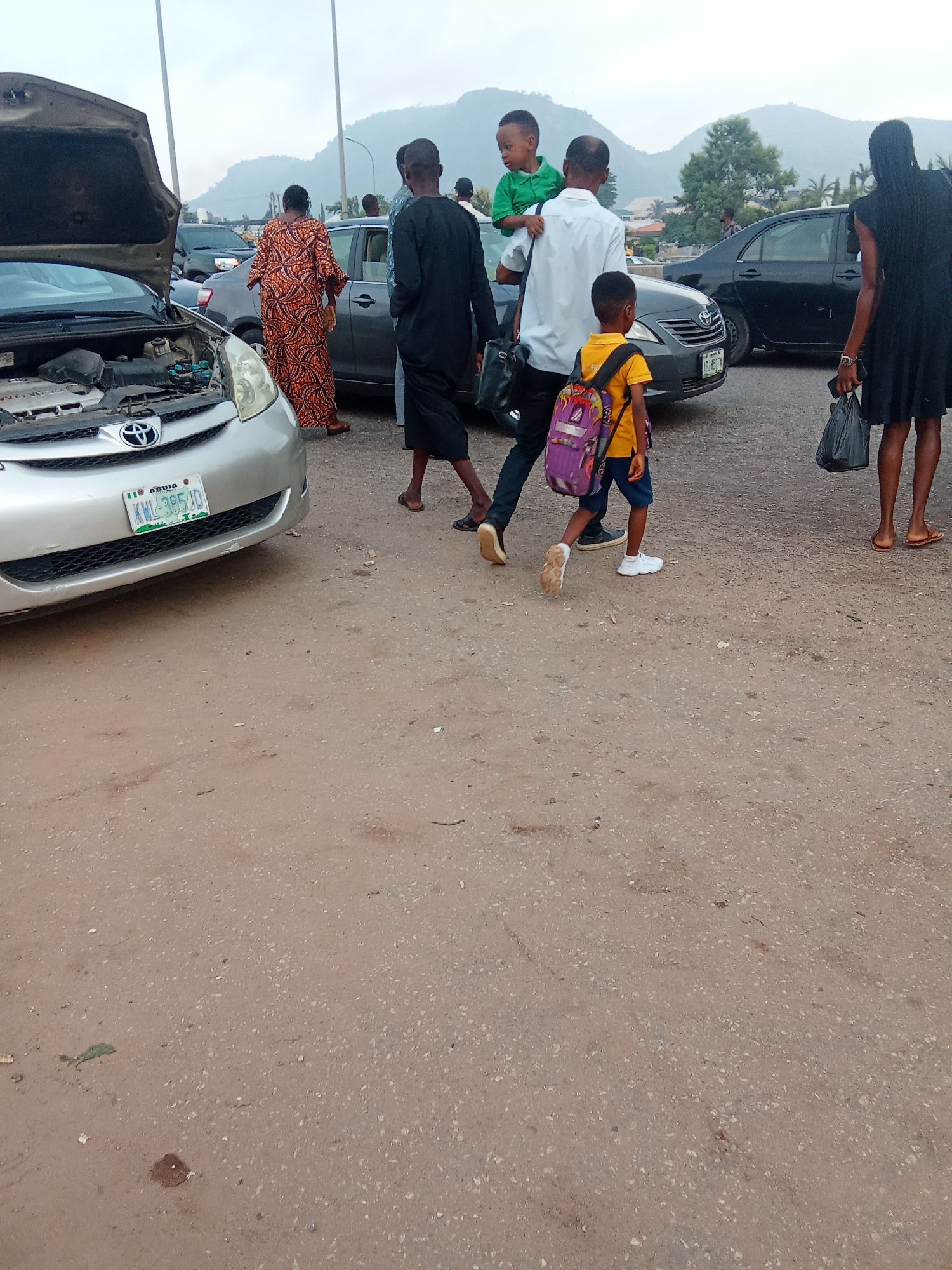 In Kagini, Nigeria, a group of people are gathered near a silver Toyota minivan with its hood open. A woman in an orange and black patterned dress stands beside the minivan. To her right, a man in a black kaftan walks away from the camera. Further right, a man in a white shirt carries a young boy on his shoulders while walking with another young boy who has a purple backpack. A woman in a black dress walks to the far right of the frame. In the background, several cars are parked, and mountains are visible. The ground is dirt and gravel.
