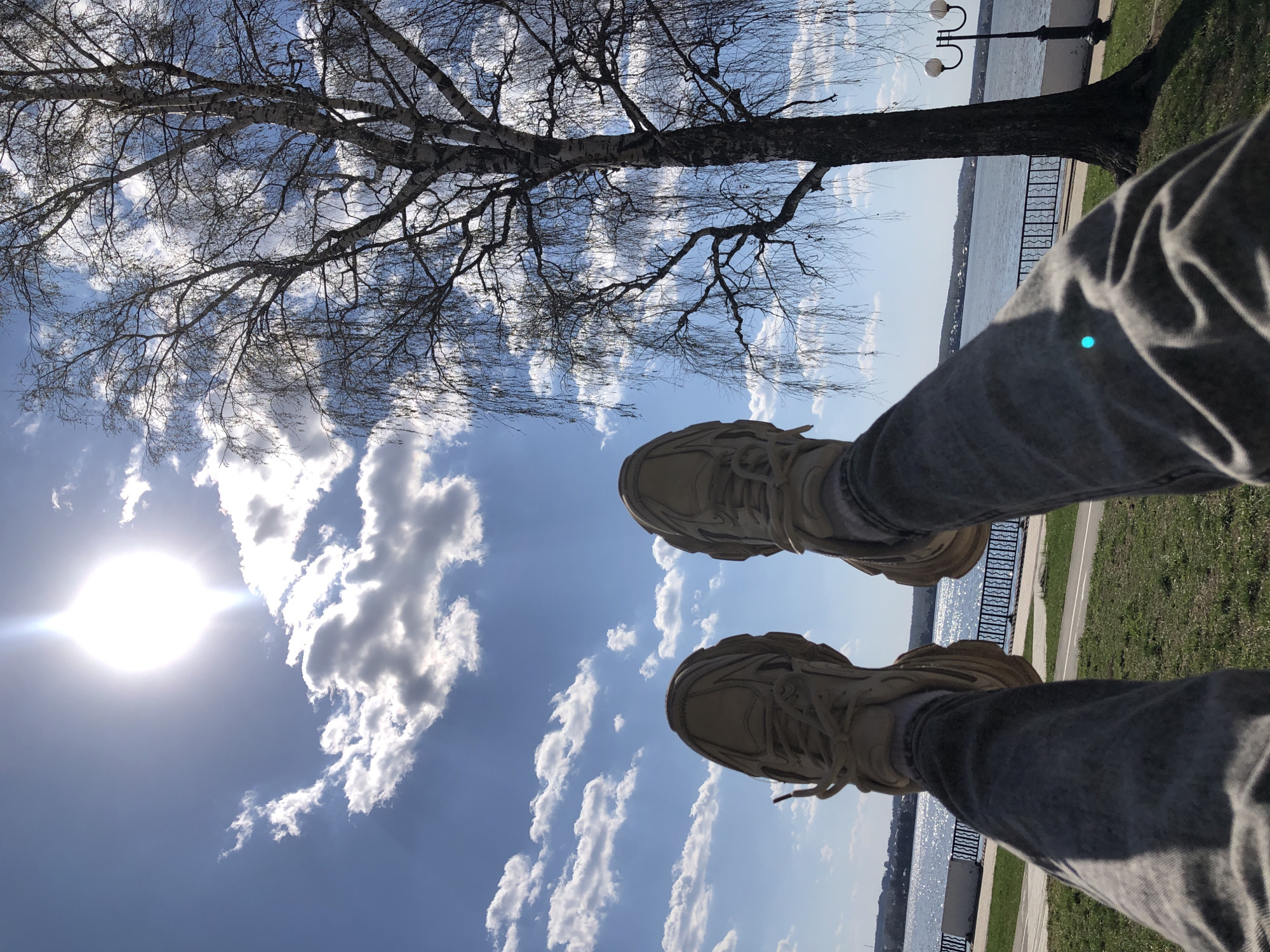 The image shows a low-angle, upward-facing view of a person's legs and shoes against a backdrop of the city of Votkinsk, Russia. The person is wearing gray jeans and light brown sneakers. The background consists of a clear sky with scattered white clouds and the bright sun. There is a bare-branched tree, a river, and a paved walkway with grass. A wrought-iron railing is visible. The scene appears to be taken in a park or waterfront area. A lamppost is visible.