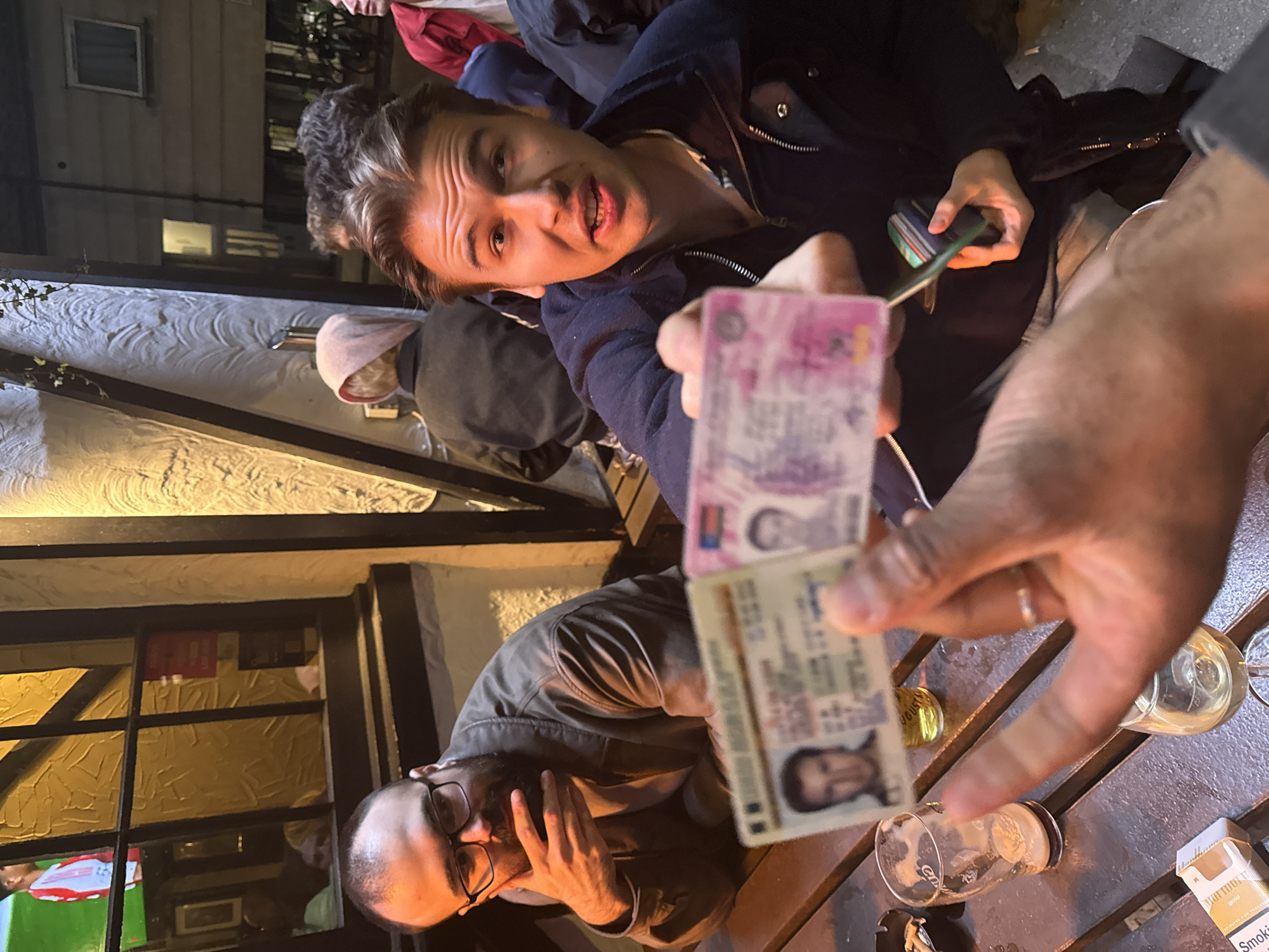 The scene appears to be set outdoors, likely a pub garden or patio in London, UK. A young man is holding up two identification cards in front of the camera, with what appears to be a wallet in his other hand. A man with glasses and a beard is seated at the table, looking down and potentially distressed or covering his face with his hand. The table holds empty glasses, a lighter and a pack of cigarettes. Another individual is partially visible behind the young man holding the ID cards, and the architecture suggests a traditional British pub setting. It is nighttime.