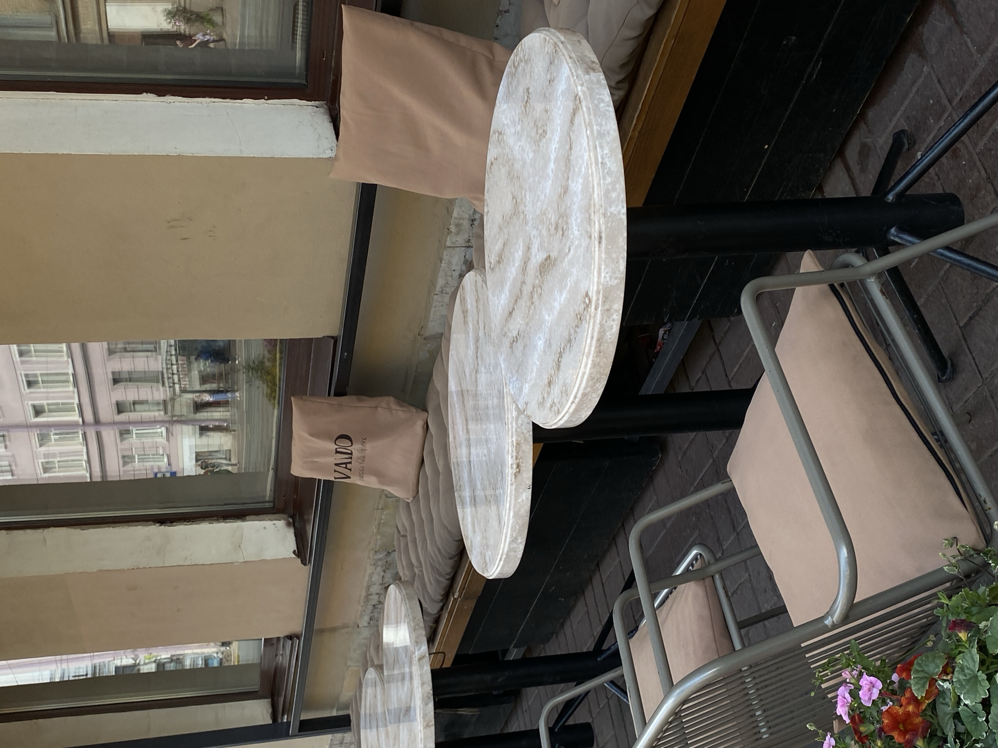 This is a medium shot, outdoor photograph of a cafe seating area in Saint Petersburg, Russia. The scene is viewed from a low angle, looking towards a building with a long window that reflects the opposite side of the street.
In the foreground, to the left, is a metal chair with a light brown cushion. Beside it, a pot of flowers with red and pink blooms is visible. Behind the chair, there are two round tables with marble-patterned tops and black metal bases. To the right of the tables is a long bench with a thick, tufted light brown cushion, and behind that, a large, light brown pillow is leaning against the wall.
The building's facade is a muted yellowish-beige, with a darker band above the window frame. The window itself has a dark brown wooden frame and reflects a street scene with several multi-story buildings opposite, featuring numerous windows and architectural details. There are blurred figures of people walking on the sidewalk across the street, and a parked car is partially visible.
The ground is paved with brick-like tiles. A brown paper bag with "VALDO" printed on it in black lettering sits on the bench, slightly to the left of the pillow. The lighting suggests it is daytime, with soft shadows. The overall atmosphere is that of a quiet, slightly overcast day in an urban setting.