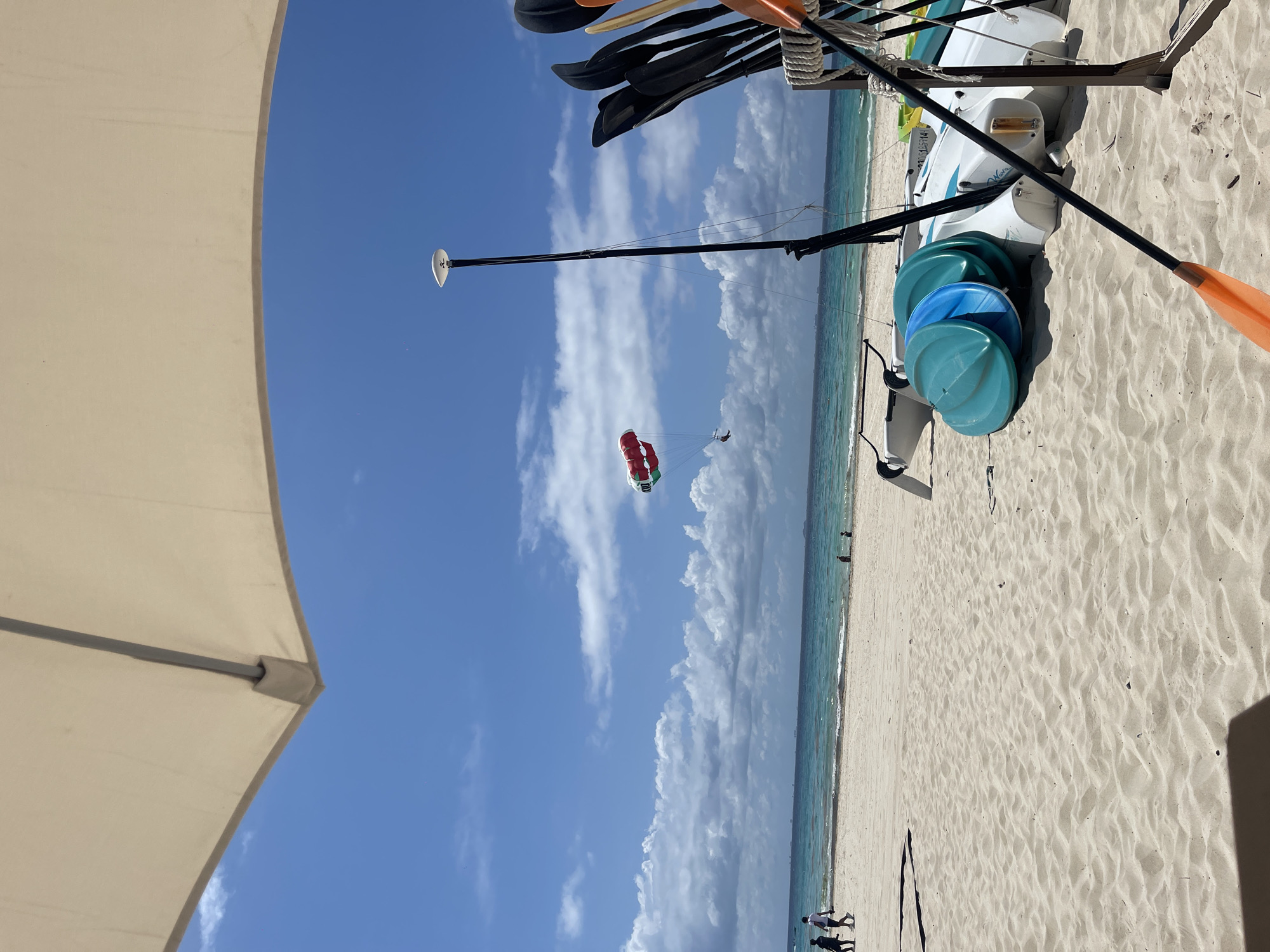 The image captures a beach scene in Playa del Carmen, Mexico. The foreground shows the edge of a partially visible white parasol. To the right, a collection of kayaks and paddles are stored on the sandy beach. In the background, the turquoise ocean meets a white sandy beach. A parasail with a red and white parachute is visible in the sky, with a person suspended beneath it. The sky is a bright blue with scattered white clouds. At the bottom of the frame, people are walking along the shore.