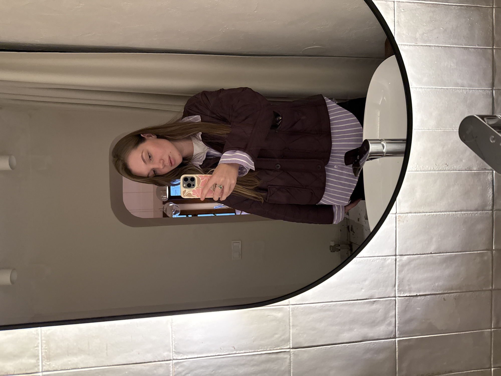 The scene is located in Belgrade, Serbia, depicting a female individual taking a mirror selfie in what appears to be a bathroom or dressing area. The subject, possessing long brown hair, is reflected from the chest up, looking downwards at the smartphone held in her left hand. She is wearing a dark purple quilted jacket over a light purple and white striped long-sleeved shirt, with a white undershirt visible at the collar. A silver ring is present on her left ring finger. The smartphone case is light-colored with a pink floral pattern.
The mirror is oval-shaped with a black frame, and its left edge emits light, suggesting integrated illumination. Reflected in the lower portion of the mirror is a white, oval-shaped countertop basin with a modern silver single-lever faucet. The actual wall surrounding the mirror on the left side is covered in vertically arranged, rectangular, light-colored tiles with a textured, slightly uneven surface.
The background reflected within the mirror reveals a room with light, neutral-colored walls. To the right of the subject's reflection, a floor-to-ceiling light-colored curtain is visible. An arched doorway in the background leads to another room, where two spherical glass pendant lights are suspended. Below the archway, a white electrical switch plate with two switches is mounted on the wall. The floor in the reflected background area appears to be in an unfinished or under-renovation state, with debris visible. A white cylindrical ceiling light fixture is also reflected in the upper part of the mirror.