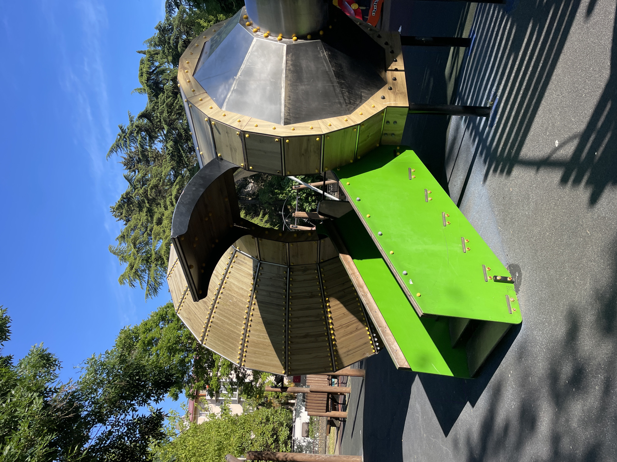 This image, taken in the city of Sochi, Russia, shows a modern playground structure under a clear blue sky. The structure is made of wood and metal, with a large cylindrical element that appears to be the central feature. One end of the cylinder is partially open, revealing a wooden interior and a ramp leading up to it. This ramp is bright green and has visible steps. The other end of the cylinder is enclosed, with large, reflective metallic panels and a clear window section. The structure is supported by dark metal legs and is situated on a dark gray, textured ground surface, likely safety surfacing for a playground. Shadows cast by the structure and surrounding trees are visible on the ground. In the background, lush green trees and a building with a red roof can be seen. The overall scene depicts a well-maintained public playground on a sunny day.