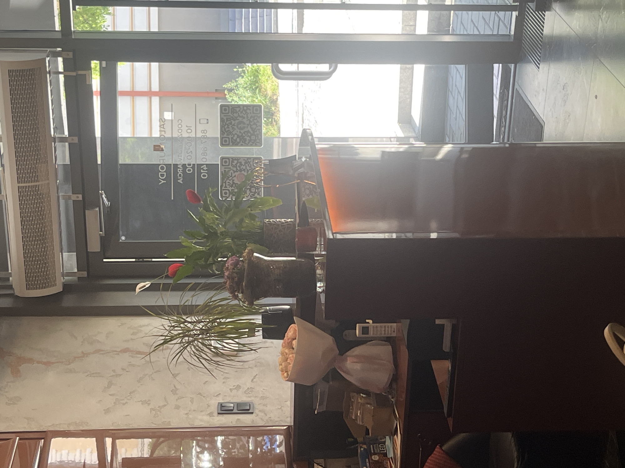The image depicts the interior of a business, likely a salon or spa, in Białystok, Poland. A reception desk is visible, adorned with a bouquet of pink flowers and a white cordless phone. Behind the desk, potted plants add a touch of greenery. The entrance features glass doors with QR codes and text, possibly indicating business hours or social media links. The overall ambiance suggests a clean and welcoming establishment.