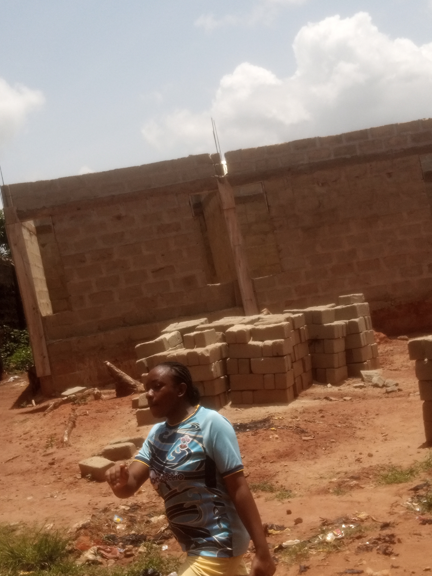 A medium shot captures a woman in a blue patterned shirt, positioned in the foreground of what appears to be a construction site in Unknown, Unknown. Her arm is extended outwards. Behind her are stacks of rectangular bricks and the beginnings of a cinder block structure. The ground is a reddish-brown dirt with sparse patches of grass and scattered debris. The sky is visible at the top of the frame, showing a mix of blue and white clouds.