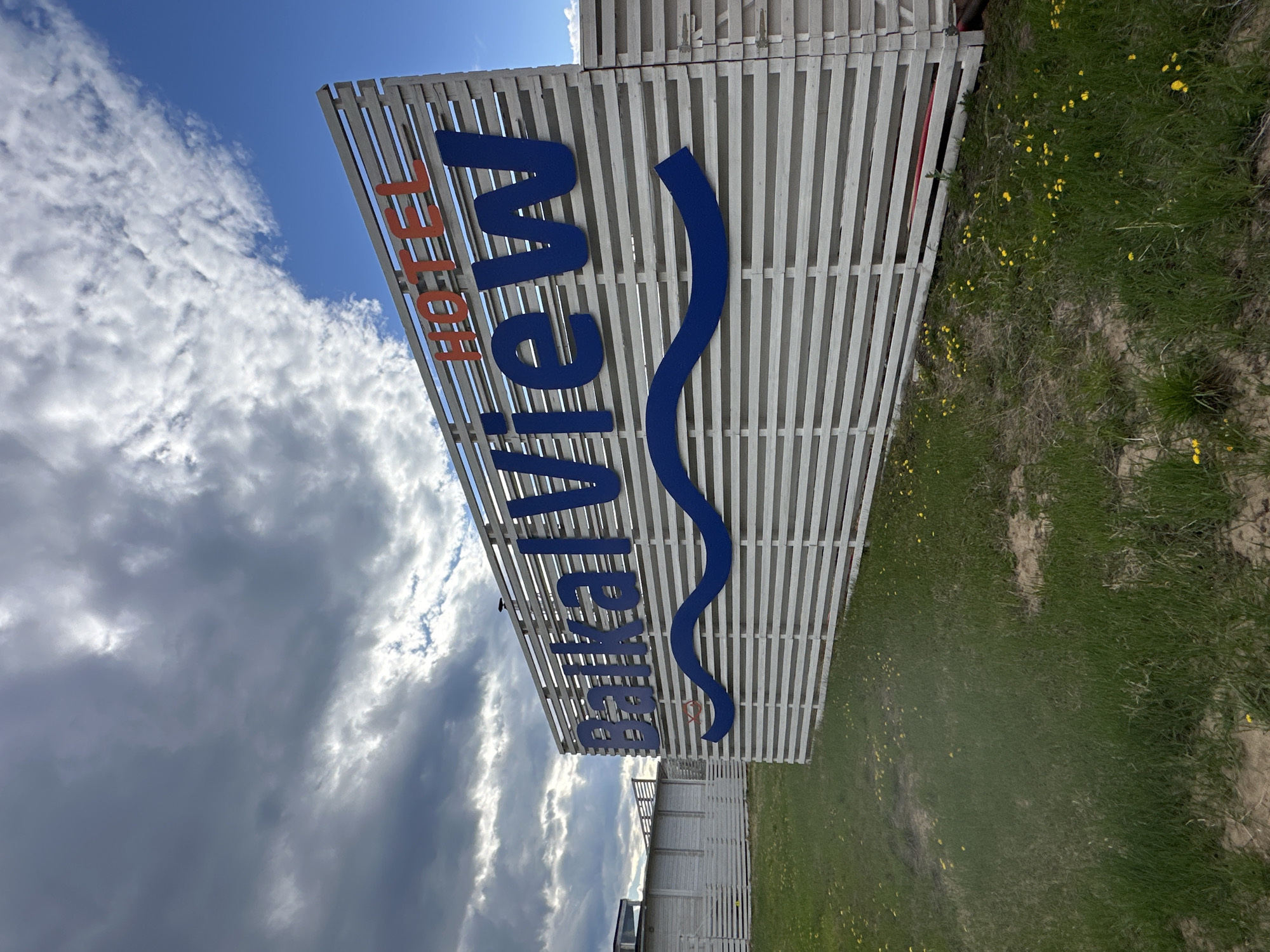 A ground-level, tilted, and full-frame photograph depicts a sign for "Baikalview HOTEL" in Khuzhir, Russia. The sign consists of white horizontal slats, with the word "HOTEL" in orange, vertical block letters. Below "HOTEL," the word "Baikalview" is written in large, blue, vertical block letters, with a wavy blue line underneath. The sky, with cumulus clouds, occupies the upper left portion of the image. Ground cover, with patches of grass and small yellow flowers, is visible on the right. A white building with horizontal slats is partially visible below the sign.