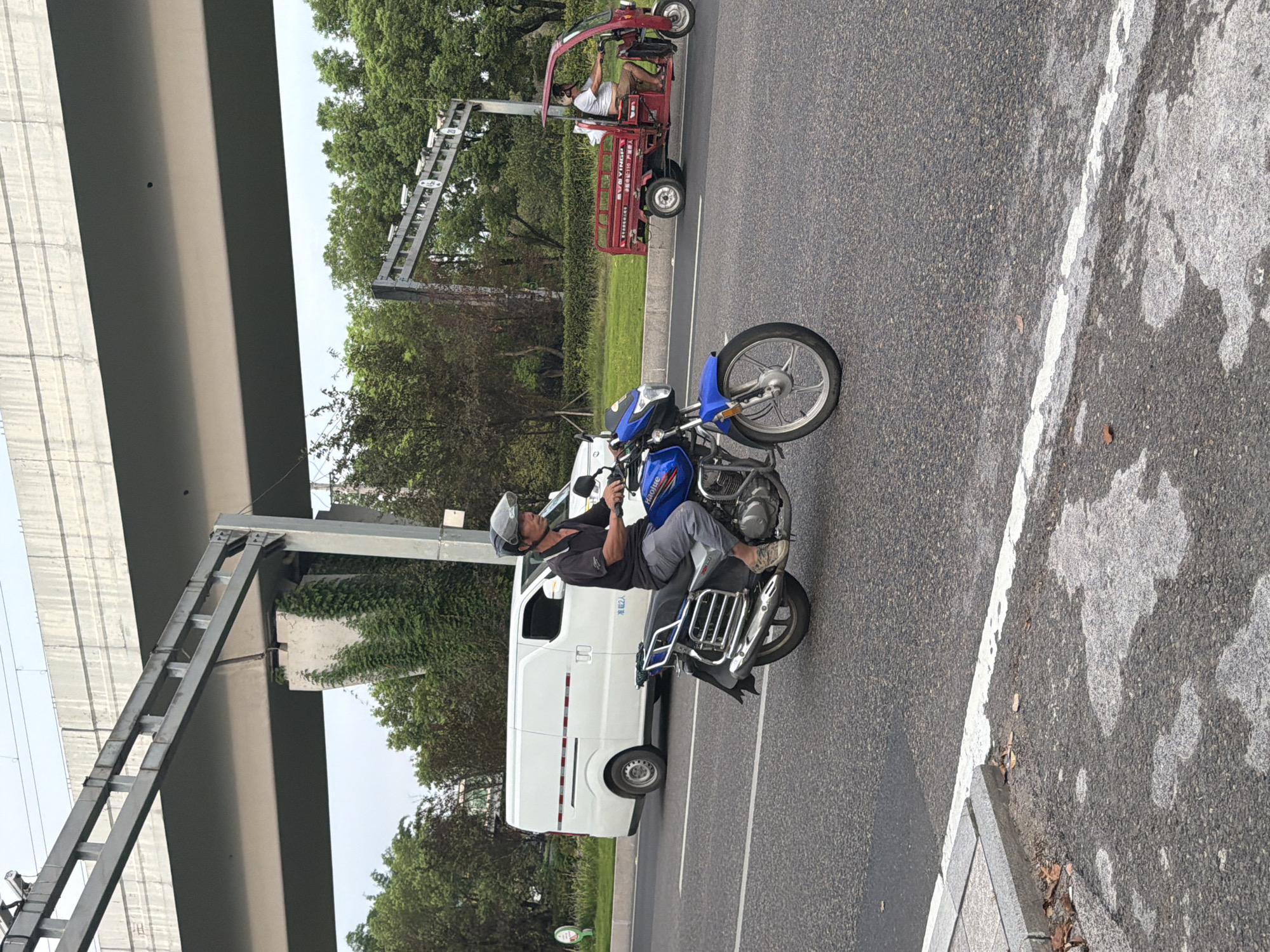 A man wearing a helmet rides a blue motorcycle on a paved road. He is dressed in a dark short-sleeved shirt and light-colored pants. A white van is parked on the side of the road behind him. Further down the road, a red three-wheeled vehicle with a person driving it is visible. The scene appears to be in an urban or suburban environment, with trees and an elevated highway structure in the background. The image was captured in Jiangjin, China.
