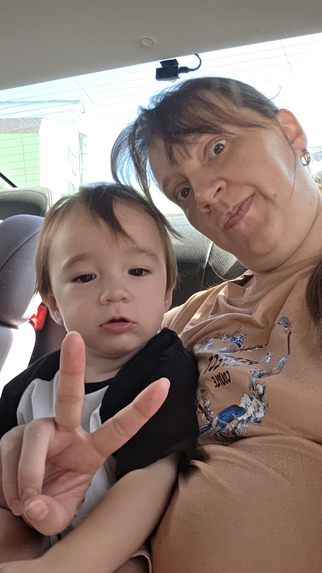 A woman and a child are in a car, likely in Dunnae-myeon, South Korea, as suggested by external context. The woman is in the foreground, looking towards the camera with a surprised expression. She is wearing a light brown t-shirt with a floral design. Next to her, a young child is also looking at the camera, holding up two fingers in a peace sign. The child is wearing a black and white shirt. The interior of the car is visible, with seats and a part of the dashboard. A small black device, possibly a dashcam, is mounted on the ceiling of the car, near the rear window. The lighting suggests it is daytime.