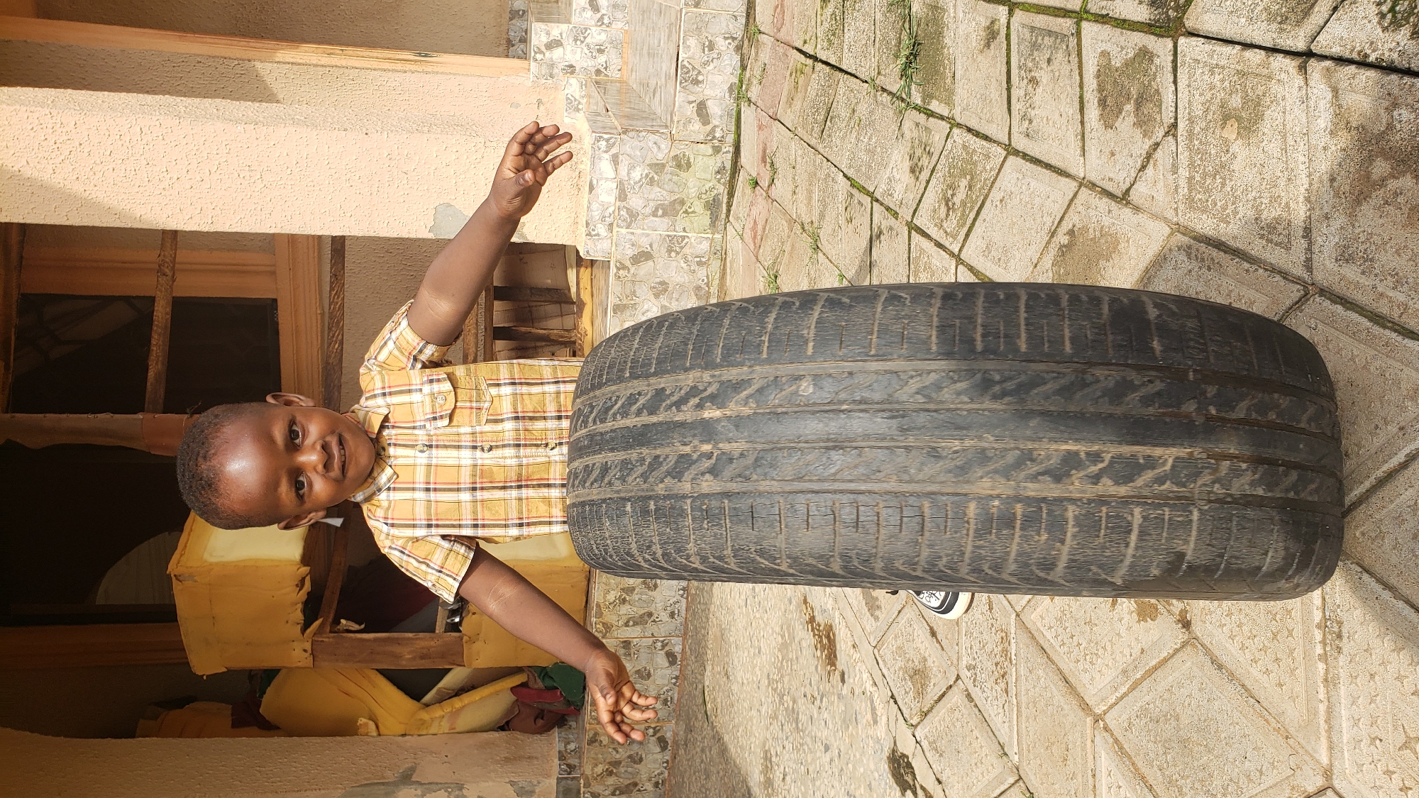 A young African boy, dressed in a short-sleeved yellow plaid shirt, stands in front of a black tire on a tiled patio. The boy has dark skin, short dark hair, and is smiling at the camera. His arms are outstretched, with his left hand slightly raised and his right hand extended towards the tire. The tire is upright and takes up a significant portion of the foreground. The background features a building with a wooden doorway and some yellow foam cushioning visible through the opening. The tiled patio is light-colored with some areas of discoloration and small green weeds growing in the cracks. The overall scene appears to be outdoors in a residential area.