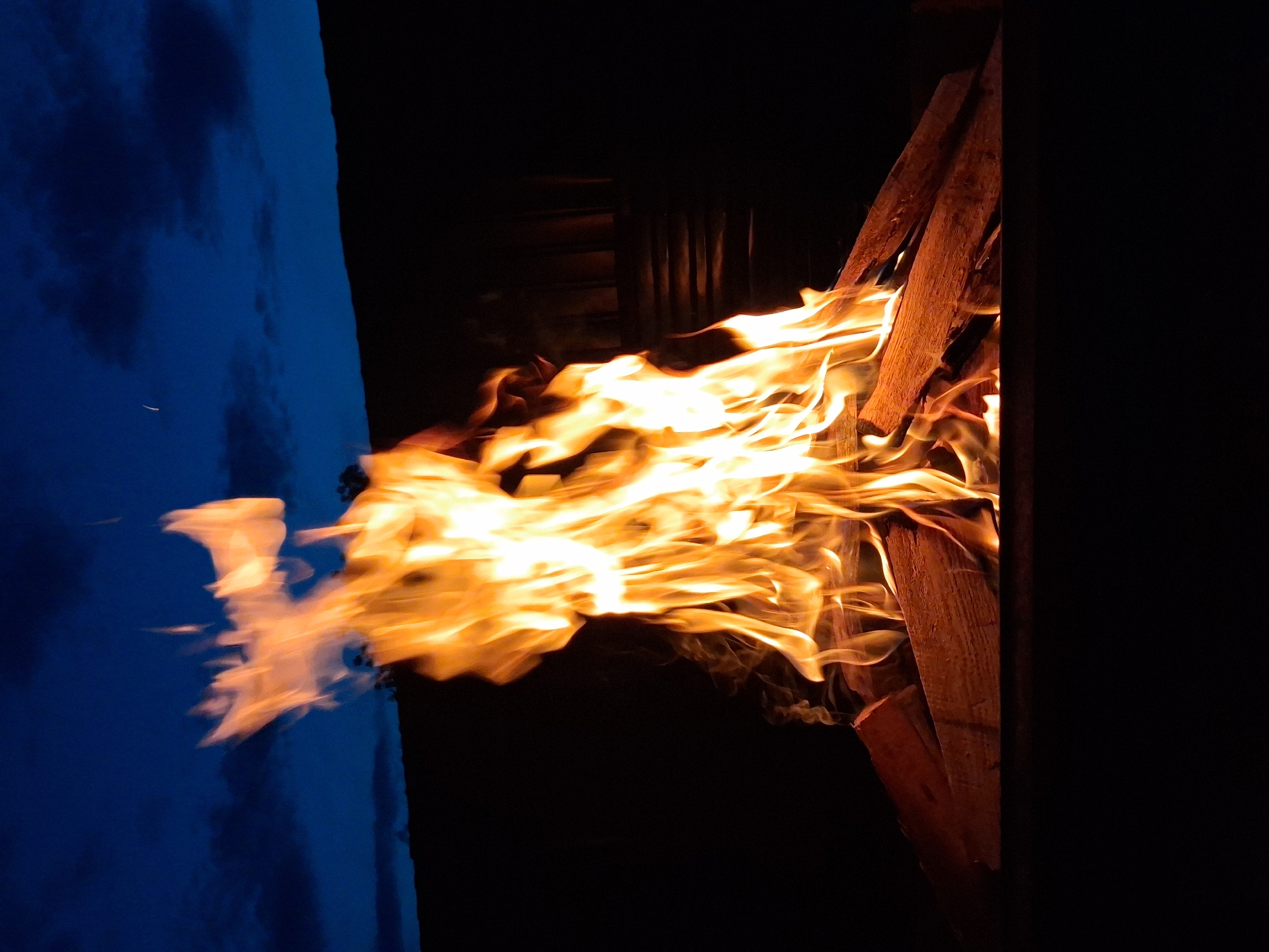A bonfire burns brightly in the darkness. The flames are a vibrant orange and yellow, casting a warm glow on the surrounding area. The fire is built on a bed of logs. In the background, a dark silhouette of a hill is visible against a deep blue night sky with scattered clouds. There is no indication of the city of Semey, Kazakhstan.