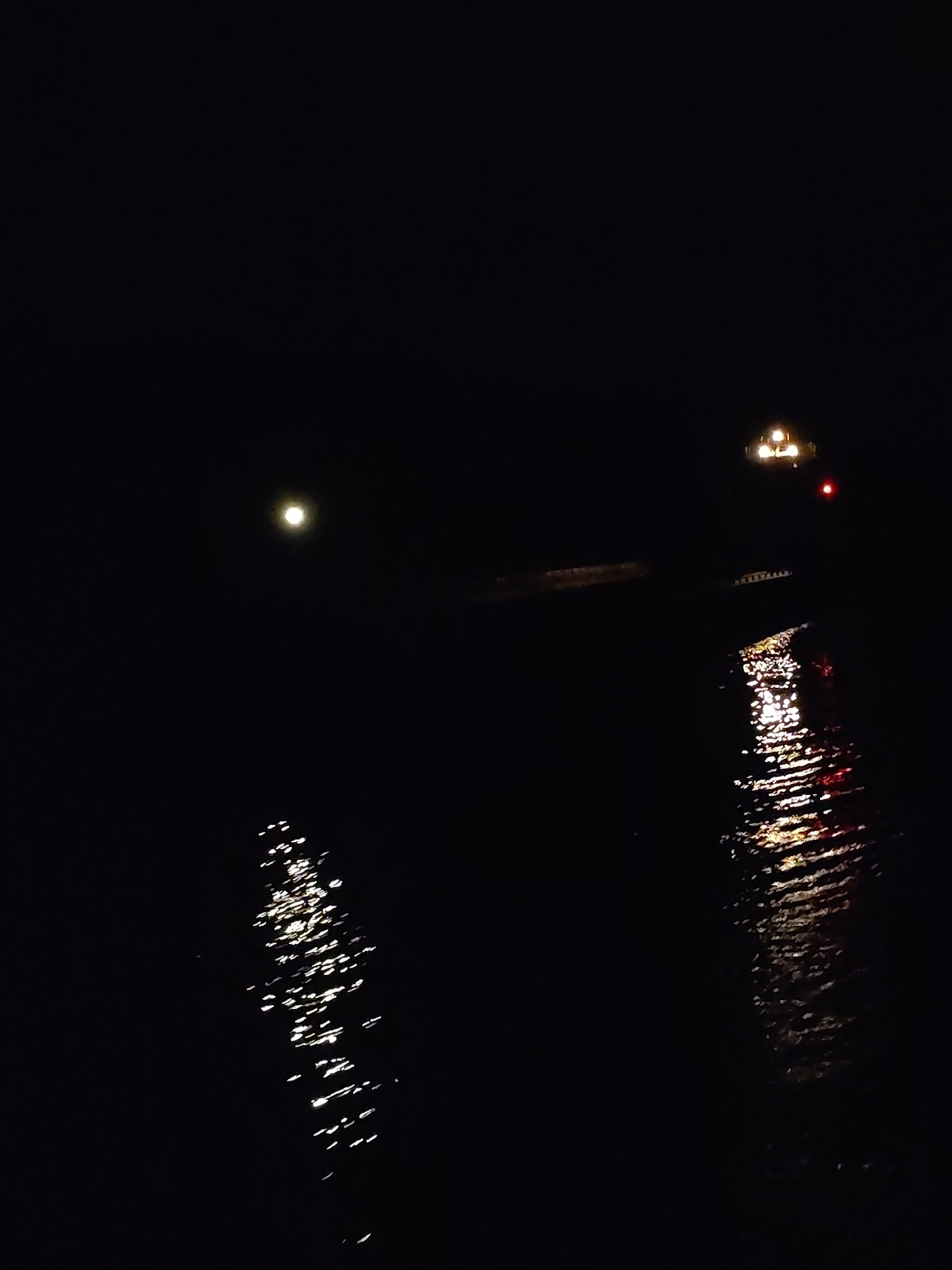 The image depicts a tranquil nighttime scene over a body of water, likely a river or lake, in Kargach, Russia. The sky and the majority of the water are enveloped in deep darkness, characteristic of night.
In the upper left portion of the frame, a distinct, bright, circular light source is visible, resembling the moon, casting a soft, ethereal glow. Below this, elongated streaks of white and silver light shimmer on the water's surface, indicating reflections from the moon or another nearby light, and suggesting gentle ripples or currents.
Further to the right, another cluster of lights breaks the darkness. Several bright yellow and white lights are prominent, appearing to originate from a distant structure or vessel, possibly a boat, a bridge, or a building on the far bank. A single red light is also visible near this cluster. These lights create vibrant, elongated reflections on the water, appearing as warm streaks of yellow, orange, and red that dance and fragment across the dark surface.
The scene is devoid of people or explicit activity, creating a peaceful and solitary atmosphere. The clear and distinct reflections suggest fair weather conditions without heavy cloud cover, fog, or rain. There is no visible text in the image. The primary subjects are the dark water, the bright celestial or artificial light sources, and their captivating reflections.