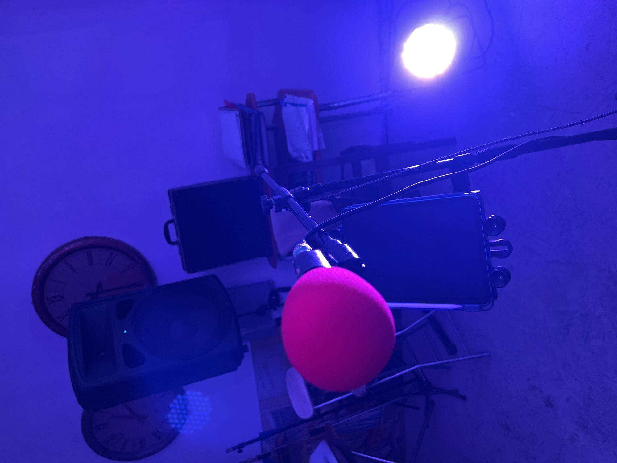 The image displays an indoor scene in Santeramo in Colle, Italy, characterized by strong blue-purple ambient lighting. A bright white circular light source is positioned on the lower right. A visible grid pattern of blue LED lights reflects onto the upper left portion of the background wall. Centrally located in the foreground is a microphone, fitted with a prominent pink foam wind screen, mounted on a stand. Directly below the microphone, a smartphone is secured to the stand. Cables extend from the stand towards the lower right. The floor is a textured, light-colored surface. Partially visible on the left are two light-seated folding chairs. In the midground, a large black PA speaker with an illuminated blue indicator light is situated on a stand. To the right of the speaker, a dark rectangular equipment case is present. Further right, a small multi-tiered cart holds stacked white binders or folders. The background wall is light-colored, featuring two large, round wall clocks with Roman numerals; the clock on the right has a reddish-brown frame. No individuals are present in the scene.
