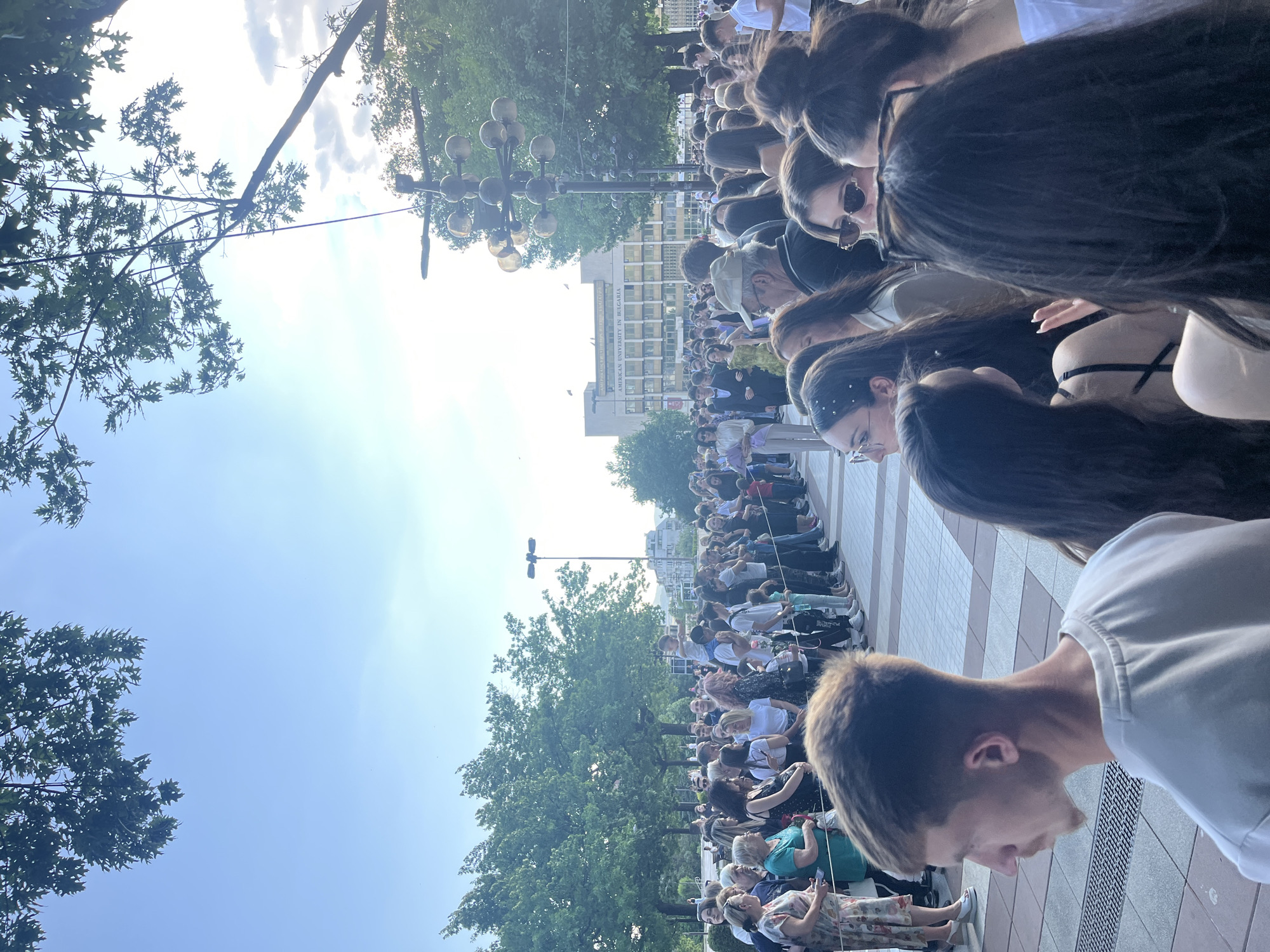 A large crowd of people is gathered in a paved plaza in Blagoevgrad, Bulgaria. The group is densely packed, facing toward the center of the image. The American University in Bulgaria is visible in the background. A row of globe-shaped light fixtures lines the plaza. The sky is blue with patches of clouds. Trees with full green foliage are present on both sides of the gathering. A person with short blonde hair and a white shirt is in the foreground, facing the crowd. Some people in the crowd are wearing white shirts.