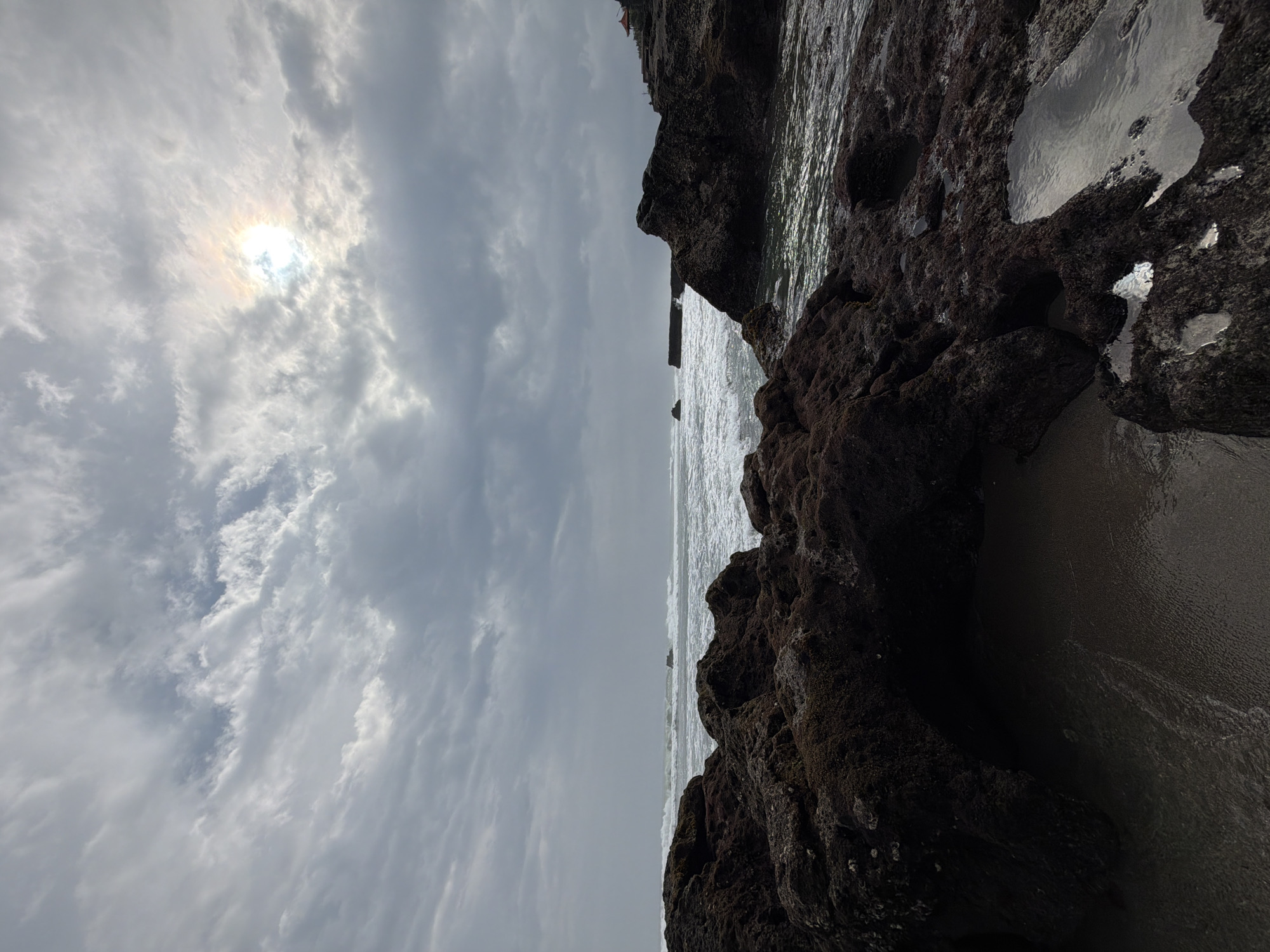 The image depicts a rocky coastline with waves crashing against the shore. The sky is overcast with some patches of sunlight breaking through the clouds. The foreground is dominated by dark, jagged rocks covered in seaweed, with small tide pools reflecting the sky. In the distance, the ocean stretches to the horizon, with white foam from the waves visible. A small red-roofed structure can be seen on the cliff edge to the right. The location is the beach in North Kuta, Bali, Indonesia, likely Tanah Lot or a nearby area given the distinctive rock formations and temple-like structure. There are no people or animals visible in the image.