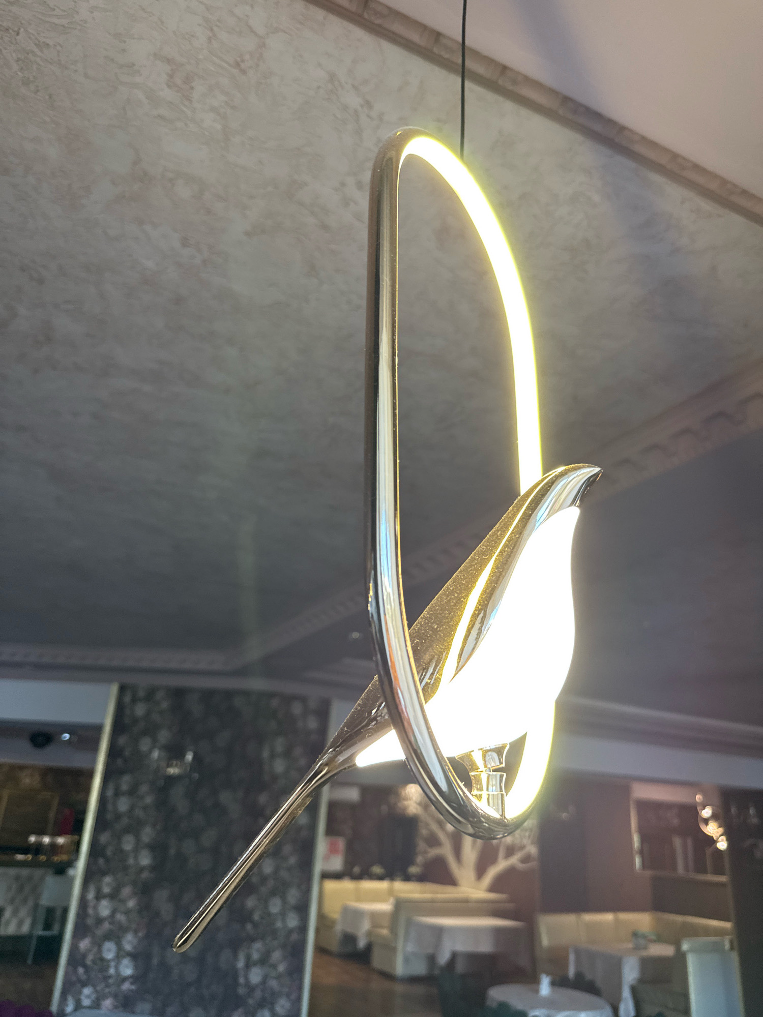 The image shows a close-up of a decorative pendant light fixture in the shape of a bird with a polished metallic body and a glowing LED ring. The background appears to be an interior space with tables and seating, suggesting a restaurant or dining area. The ceiling has a textured, marbled finish with ornate trim. The lighting is warm, with the LED ring casting a bright yellow-white glow. The bird's beak and tail are elongated, and its body is smooth and reflective. Some dust particles are visible on the fixture. The location is identified as Torzhok, Russia.