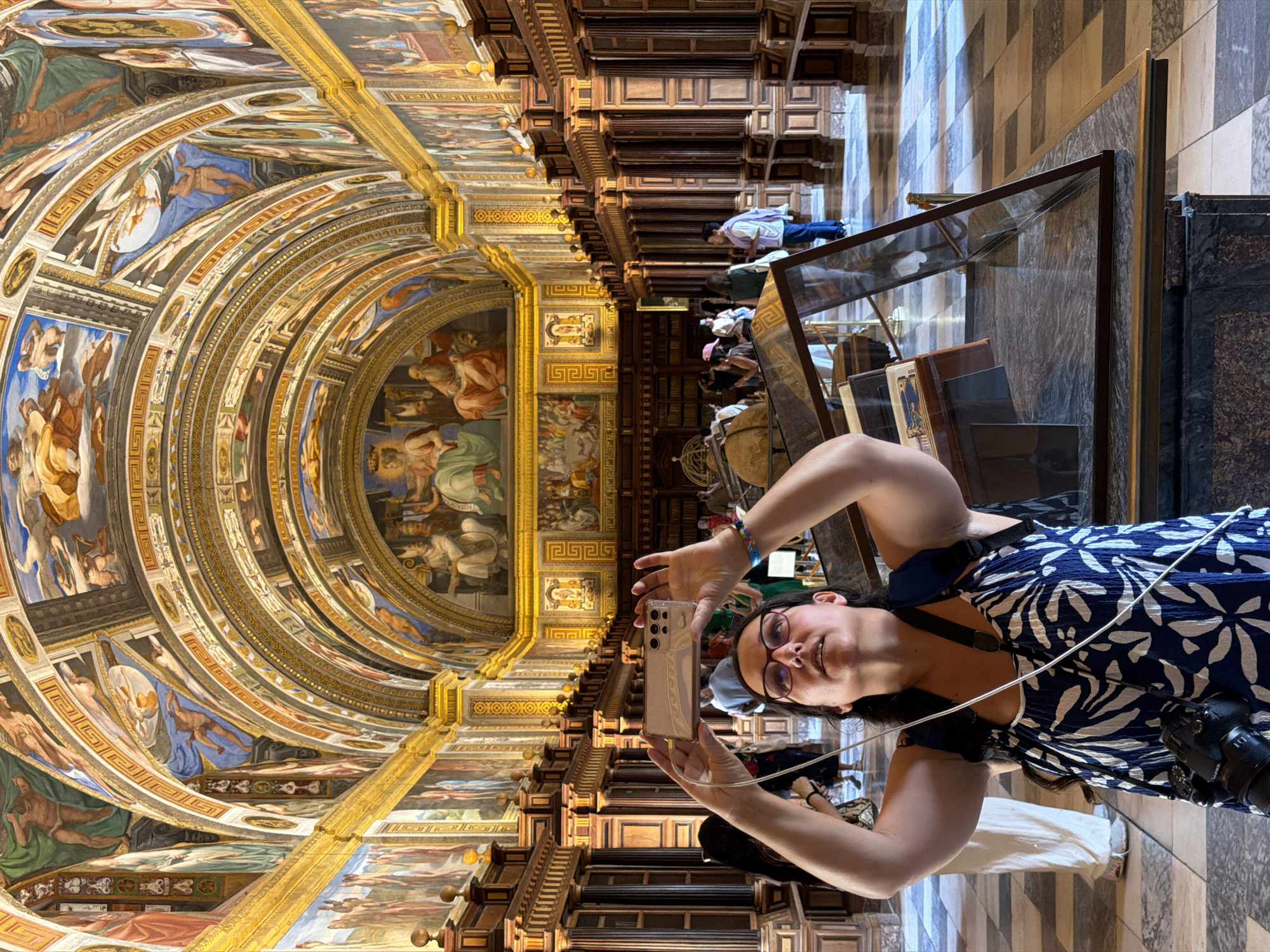 The image shows a woman taking a picture of the ornate ceiling frescoes in the Royal Library of San Lorenzo de El Escorial, Spain. The woman is in the foreground, holding a smartphone in landscape orientation. She is wearing glasses and a blue and white floral dress, with a camera around her neck. The library's interior is characterized by high, vaulted ceilings adorned with vibrant frescoes, and wooden bookshelves lining the walls. Other visitors are visible in the background, as well as display cases containing artifacts. The floor is tiled in a checkered pattern. The lighting is bright, illuminating the rich details of the architecture and artwork.