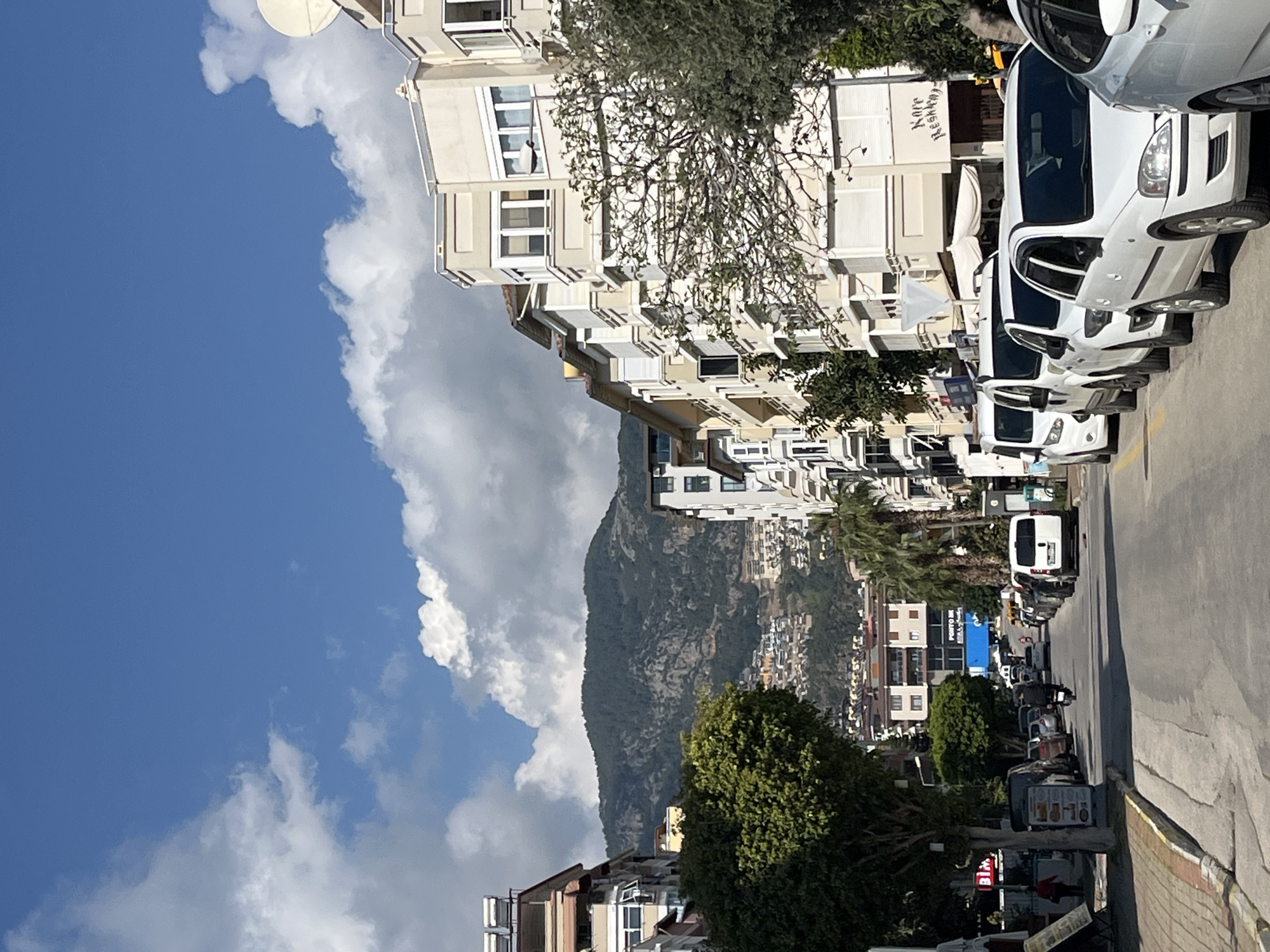 The image shows a street scene in Alanya, Turkey. On the right side of the frame, multiple white cars are parked along the road. Buildings line the left side of the street, with apartments or residential structures visible. A mountain rises in the background, partially obscured by clouds in the blue sky. Vegetation, including trees, is interspersed throughout the scene. A street is visible. In the distance, other buildings are seen on the mountain side. The sky is clear and blue.