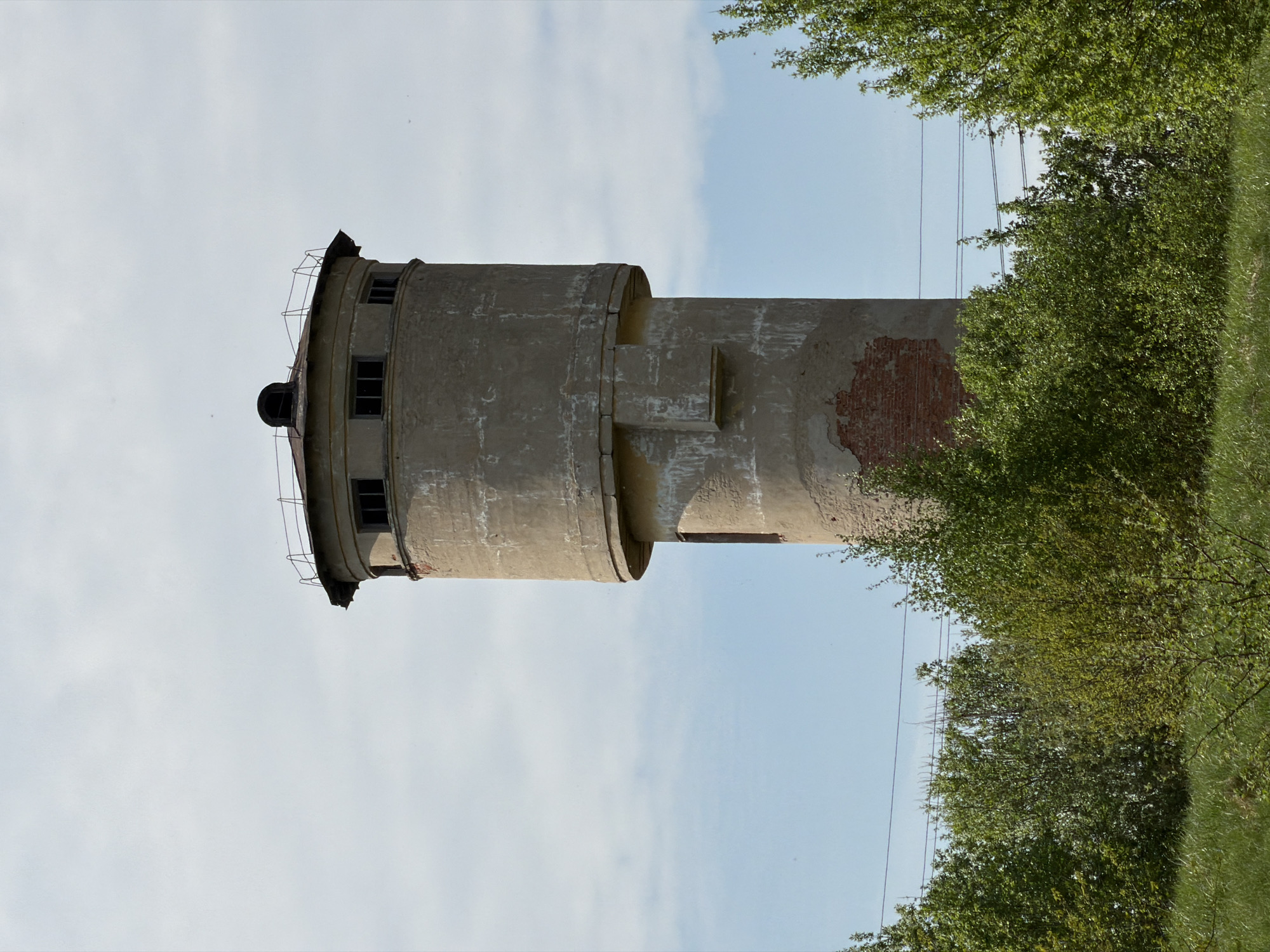 An outdoor shot features a concrete water tower with some brickwork visible, set against a backdrop of a pale blue sky with scattered clouds. The tower appears cylindrical, with a wider section near the top that houses small windows and a small domed roof. A rusted railing encircles the top section. Concrete is showing signs of wear and tear. To the right of the tower, green foliage partially obscures the structure, creating a natural frame. Power lines are visible running vertically along the right side of the image, threading through the branches of the trees. According to information provided, the location of the image is Tokkari, Russia.