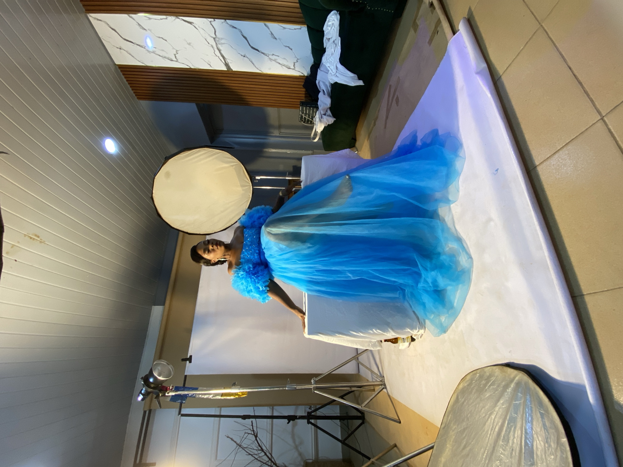 In Bukuru, Nigeria, a woman is seated on a white-covered table in a studio. She wears a bright blue, off-the-shoulder gown with voluminous puffy sleeves and a flowing skirt. The studio setting includes professional lighting equipment, including a large softbox to the right and lighting stands to the left. A white backdrop is positioned behind the woman, and the floor appears to be tiled. A green sofa with some draped clothing is visible in the background, along with a decorative wall panel. The overall atmosphere suggests a professional photoshoot in progress.