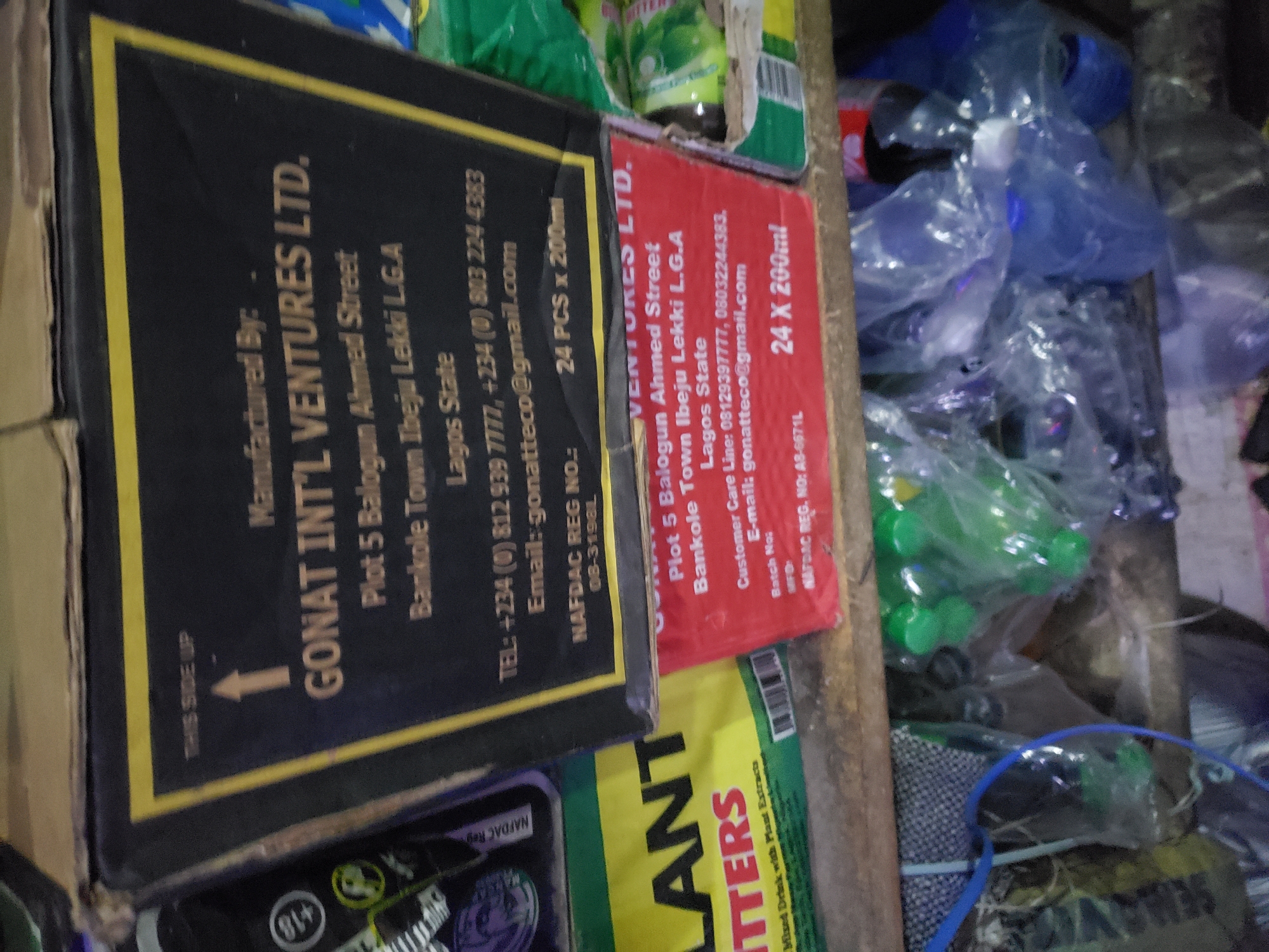 A close-up shot captures a shelf with various items in what appears to be a shop in Jos, Nigeria. The focus is on a black cardboard box with a yellow border that has text printed on it. The box is oriented so that "THIS SIDE UP" is visible at the top, with an arrow pointing upwards. Below this, it reads "Manufactured By: GONAT INT'L VENTURES LTD." Further details on the box include the company's address: "Plot 5 Balogun Ahmed Street, Bankole Town Ibeju Lekki L.G.A, Lagos State." Contact information is also present: "TEL: +234 (0) 812 939 7777, +234 (0) 803 224 4383" and "Email: gonatteco@gmail.com". A "NAFDAC REG NO.: 08-3198L" is also visible. The box indicates the contents are "24 PCS x 200ml".
Below this box, a red cardboard box is partially visible, also with text indicating "VENTURES LTD." and an address that mirrors the one on the black box. This red box also shows "24 X 200ml" and a "NAFDAC REG. NO: AB-6671L".
To the left of these boxes, a portion of a green cardboard box is seen with the word "LANT" and below it "ITTERS" printed in large black letters. Underneath this, smaller text reads "Mixed Drink with Plant Extracts."
The shelf itself appears to be made of wood and is cluttered. Behind and below the boxes, various plastic bags filled with items are visible. Some of these bags contain green spherical objects, possibly bottle caps or small toys, and other transparent bags hold clear or colored liquids, likely beverages. The overall impression is of a retail environment, possibly a small store or market stall.