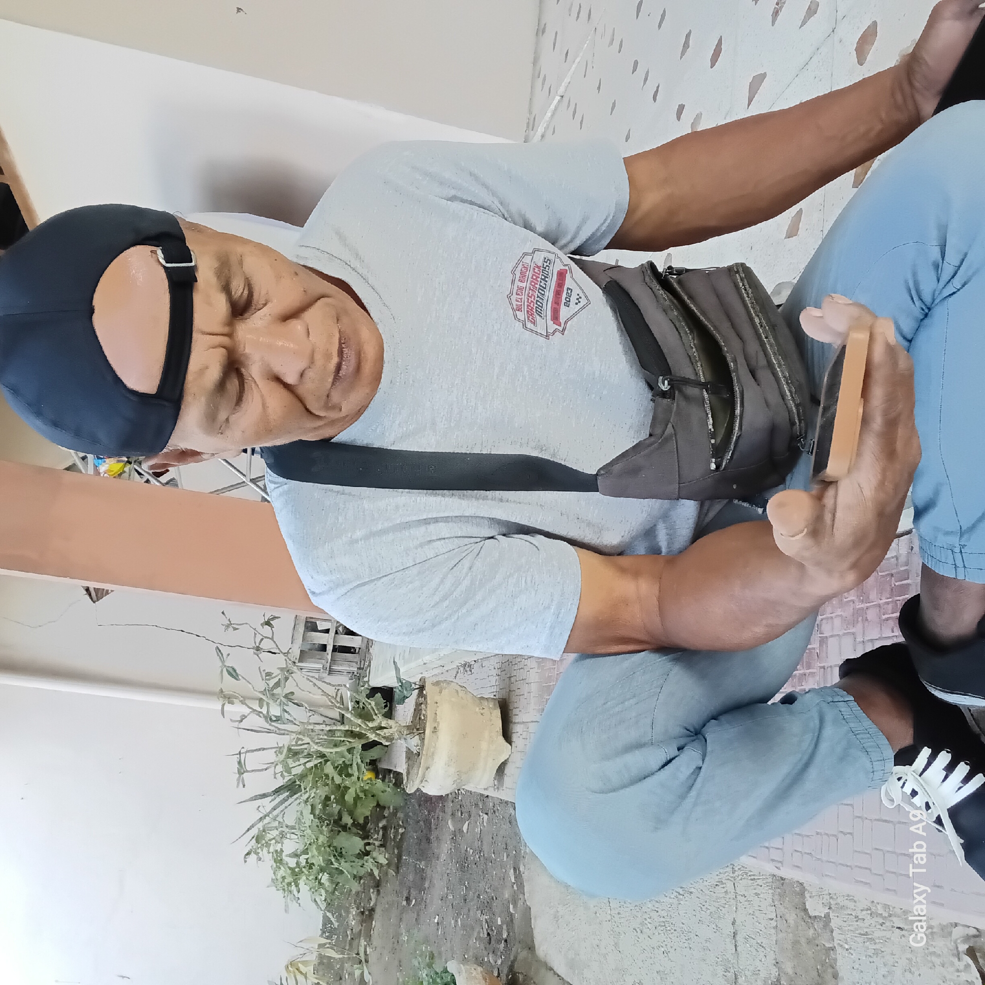 A man wearing a grey t-shirt, blue pants, and a black baseball cap is kneeling on a tiled surface outdoors. He is holding a small orange object in his right hand and looking down at it. A brown bag is slung across his chest. In the background, there are potted plants and a light-colored wall. The scene appears to be in an outdoor residential area in Sambirejo, Indonesia.