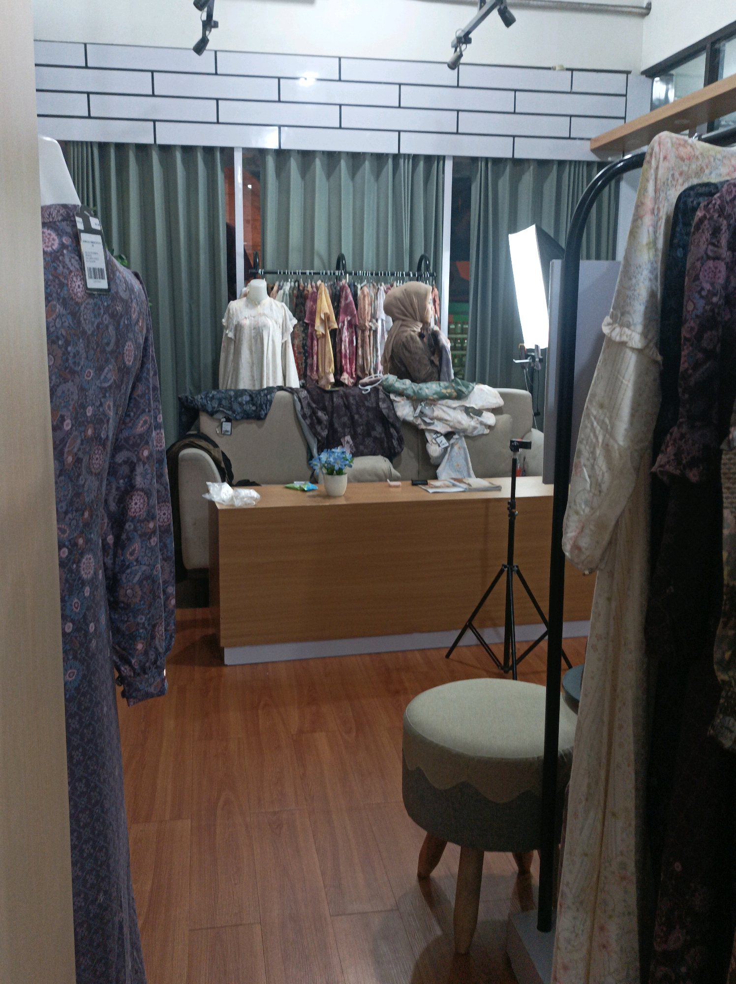 The image depicts an indoor scene, likely a retail or studio space, in Blimbing, Indonesia. In the foreground, a mannequin is draped with a patterned garment, partially obscuring a view of a wooden floor. To the right, a rack of clothing is visible, with several garments hanging. In the background, a beige couch is positioned in front of a green curtain. A person wearing a headscarf is seated on the couch, appearing to be arranging clothes or items. Adjacent to the couch is a wooden table with a small potted plant containing blue flowers and some plastic items. A professional lighting setup, including a large rectangular light panel on a stand and a tripod with a camera mount, is present in the middle of the room, suggesting the space is used for photography or videography. The overall environment appears to be a tidy, well-lit space with elements of both commercial display and production.