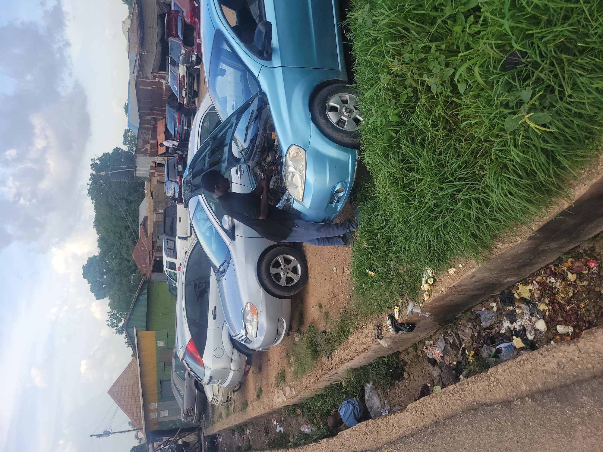 A man is working on the engine of a light blue car with its hood open, parked on a dirt road in Jos, Nigeria. Several other cars are parked around it, some with their hoods open as well. The scene appears to be a makeshift auto repair shop or a parking lot with various vehicles. A concrete drainage channel runs alongside the road, filled with trash. In the background, there are buildings with corrugated metal roofs and some trees. The sky is overcast with clouds.