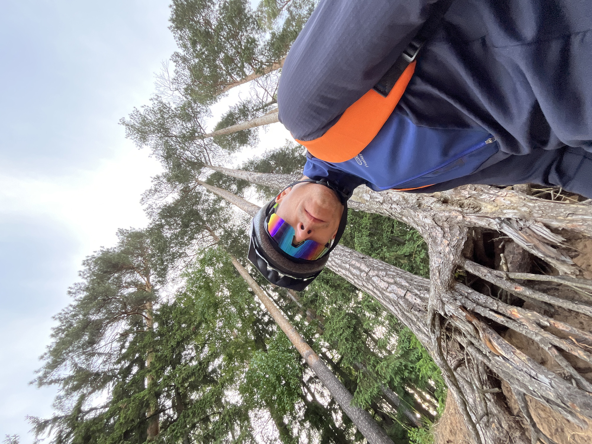 A low-angle, selfie-style photograph shows a person wearing a black and gray helmet and iridescent goggles. They are wearing a navy blue and blue jacket with an orange strap across the chest. The person is standing in a forest with tall pine trees reaching upwards into a cloudy sky. The camera is angled upwards, making the person appear to be looking down at the viewer. The trees and foliage suggest a wooded area. Part of a fallen tree or root structure is visible in the bottom right corner. The photograph was taken in the city of Chaikovsky, Russia.