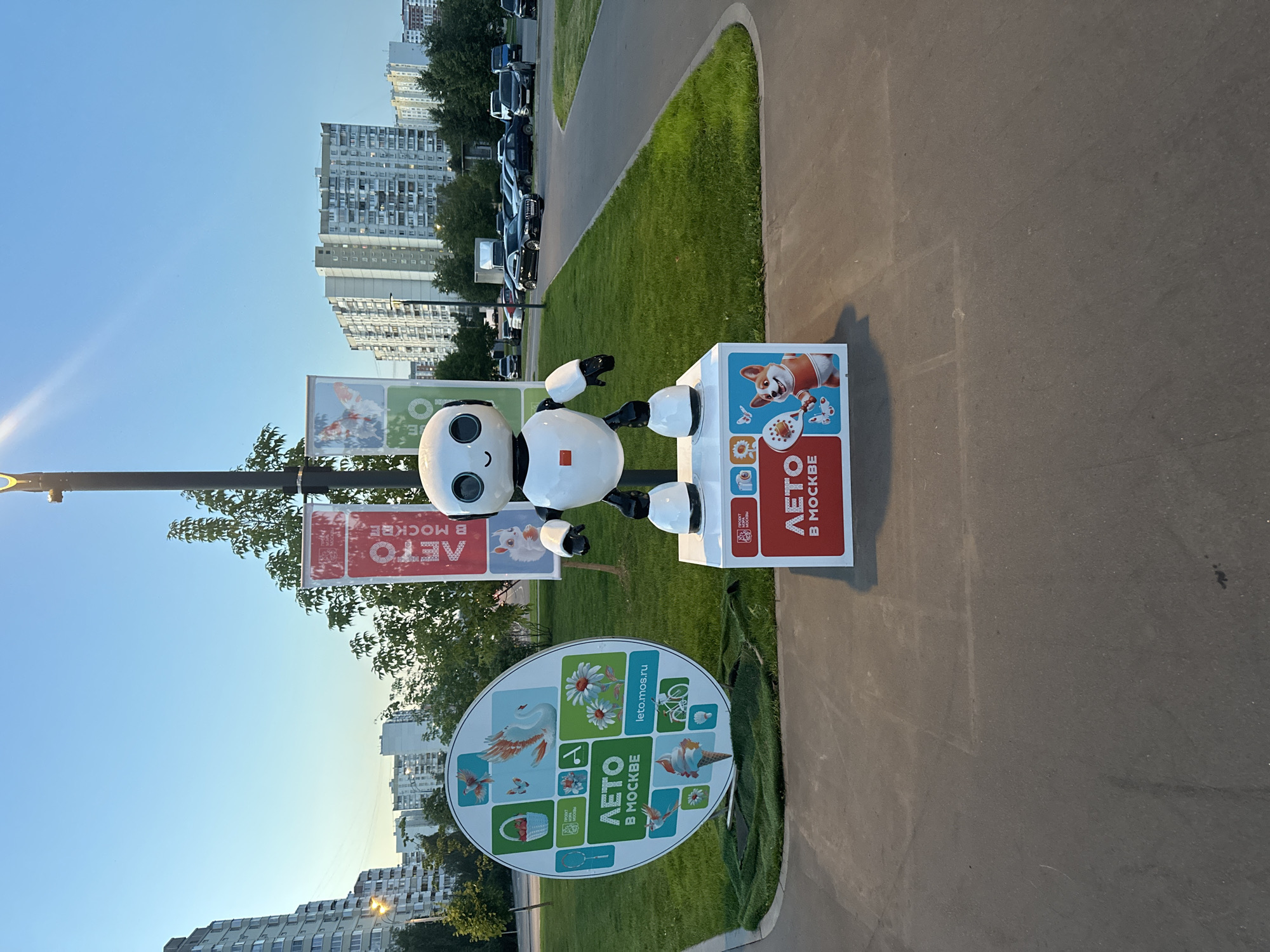 A white, anthropomorphic robot with a black head and large, circular eyes stands on a white pedestal in an outdoor urban park. The robot is decorated with red accents, including a square on its chest. The pedestal it stands on is decorated with graphics, including the text "ЛЕТО В МОСКВЕ" (Summer in Moscow) and an illustration of a dog holding a tennis racket.
To the left of the robot is a large, circular sign with similar graphics, also featuring the text "ЛЕТО В МОСКВЕ" and various summer-themed images like a basket of strawberries, a swan, flowers, and an ice cream cone. The website "leto.mos.ru" is also visible on this sign.
Behind the robot and signs, banners with the same "ЛЕТО В МОСКВЕ" text hang from a lamppost. The park setting includes a well-maintained green lawn, a paved walkway, and trees. In the background, several multi-story apartment buildings characteristic of Moscow's residential architecture are visible. Cars are parked and driving on a road in the distance. The sky is clear and blue, suggesting it is daytime, likely late afternoon or early evening due to the lighting. The overall scene suggests a public art installation or promotional display for a summer event in Moscow.