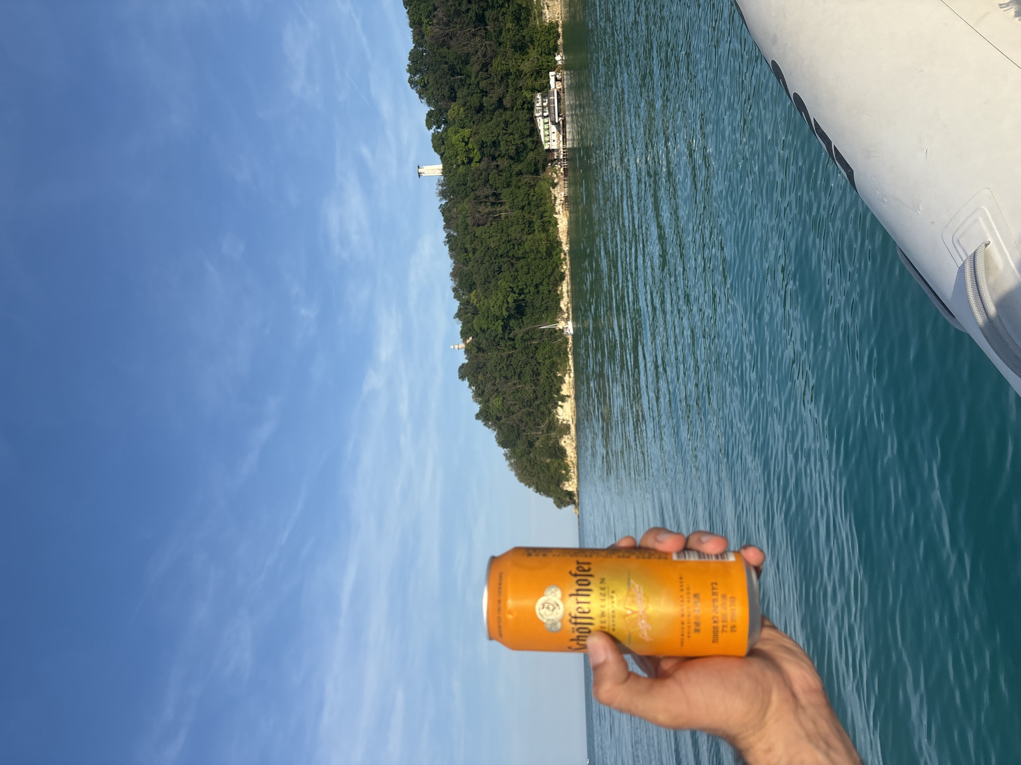 A first-person perspective image captures a scene from a boat on a body of water, facing a forested coastline under a blue sky. In the immediate foreground, a hand with light-to-medium skin tone holds an orange "Schöfferhofer" beer can, labeled "HEFEWEIZEN GRAPEFRUIT," with Asian characters visible at the bottom. The can is held upright, partially obscuring the lower left portion of the view. The water, a clear turquoise-blue, exhibits slight ripples.
To the right in the foreground, the white, ribbed side of an inflatable boat, possibly a Rigid Inflatable Boat (RIB), is visible, including a gray rope handle and a partial black inscription "ILO" or "ZODIAC".
In the middle ground, a heavily forested cliff or hill rises from the water, displaying lighter-colored rock formations at its base. On the left side of the hill, a slender white lighthouse structure is discernible. Further to the right on the hilltop, a tall, white, square-shaped tower is visible. At the base of the cliff on the right, a multi-story, light-colored building complex, built partly over the water, is present. A small white sailboat is moored or sailing near the base of the cliff on the left, slightly to the right of the leftmost lighthouse. The sky above is clear blue with sparse, thin white cirrus clouds. The location is in the city of Unknown, Unknown.