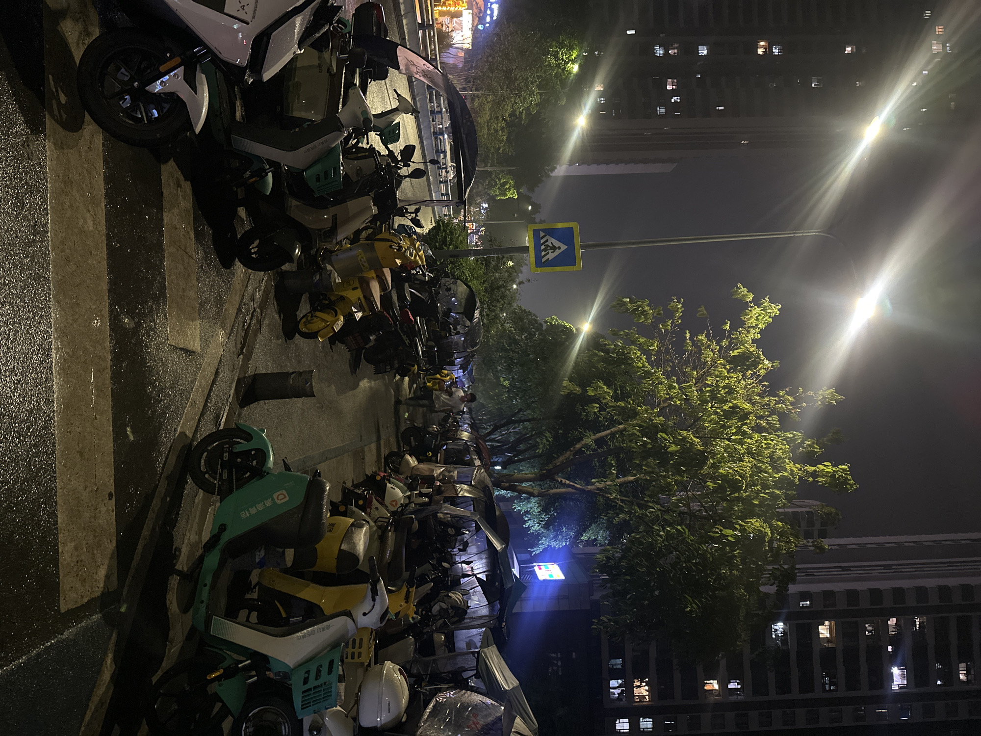 The image depicts a street in Chongqing High-tech Zone, China, at night. Numerous parked electric scooters line the street. A crosswalk is visible in the foreground. Streetlights illuminate the scene. A pedestrian crossing sign is present. Tall buildings are visible in the background, with illuminated windows. Trees are scattered along the street. The ground appears wet, possibly from rain. No people are visible in the image.