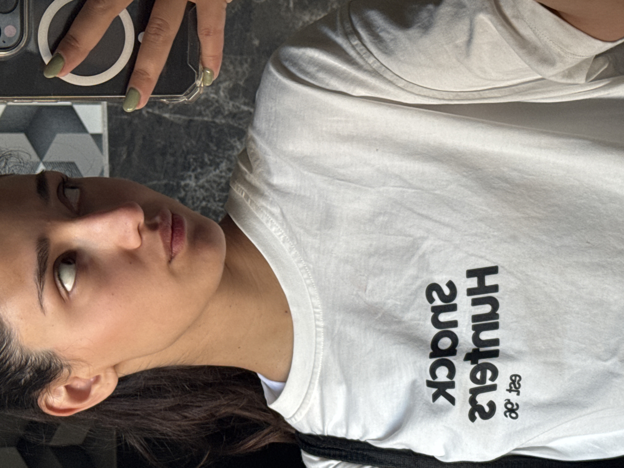 A young woman is shown from the chest up. She is holding a phone in her left hand. She is wearing a white t-shirt with black lettering that reads "Hustle snack est. '89". She appears to be inside. The exact location within the city of Bar, Montenegro is not determinable from the image.
