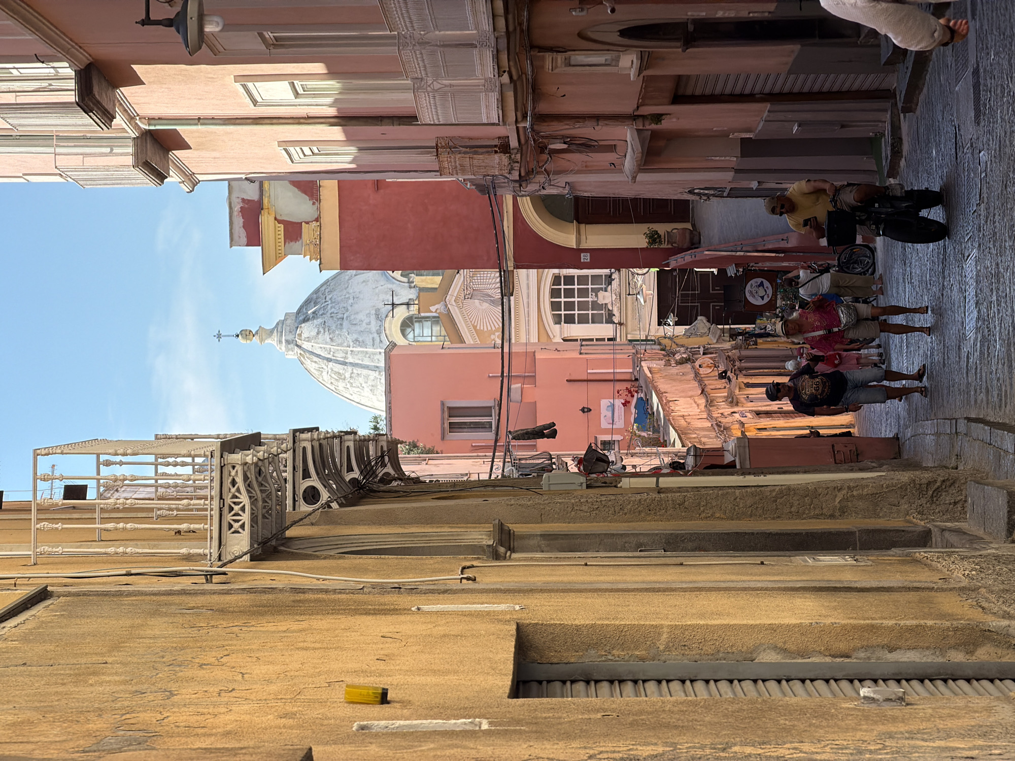 This is a vertical street scene in Procida, Italy. The foreground is a golden-hued textured building rooftop with railing and a yellow object. In the midground, a narrow street runs between colorful buildings painted in shades of pink, red, and yellow. A dome of a church is visible in the distance. Several pedestrians are walking down the street, and a man is riding a scooter. The sky is light blue and clear.