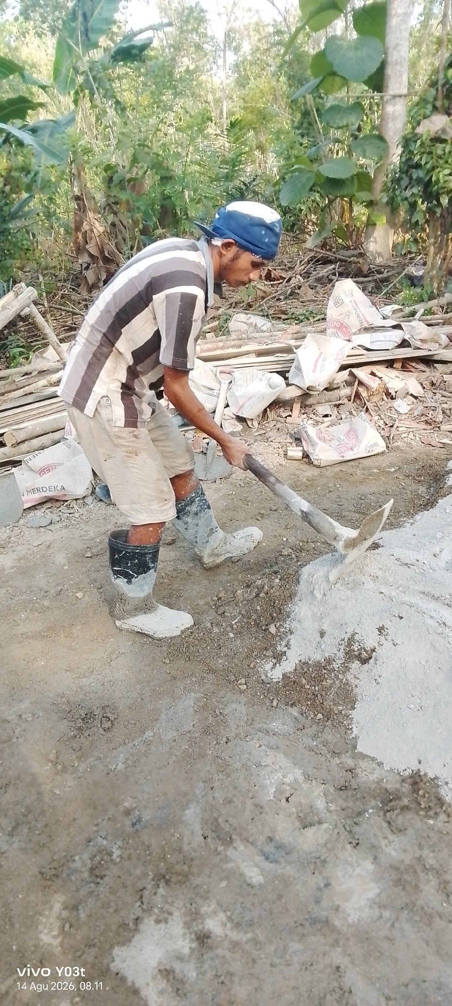 In Woiha, Indonesia, a man is actively engaged in mixing cement outdoors. He is wearing a blue and white striped t-shirt, beige shorts, and tall black rubber boots that are speckled with white cement. A blue and white bandana is tied around his head. He is holding a pickaxe with a wooden handle, using it to mix a pile of grey cement and aggregate. Behind him, several white sacks, some with red lettering that reads "MERDEKA," are stacked, along with pieces of wood and other construction materials. The environment appears to be a rural or construction site, with lush green foliage and trees visible in the background. The ground is covered in dirt and cement dust. The image is a vertical shot, captured with a phone camera, as indicated by the "vivo Y03t 14 Agu 2026, 08.11" watermark at the bottom left corner.